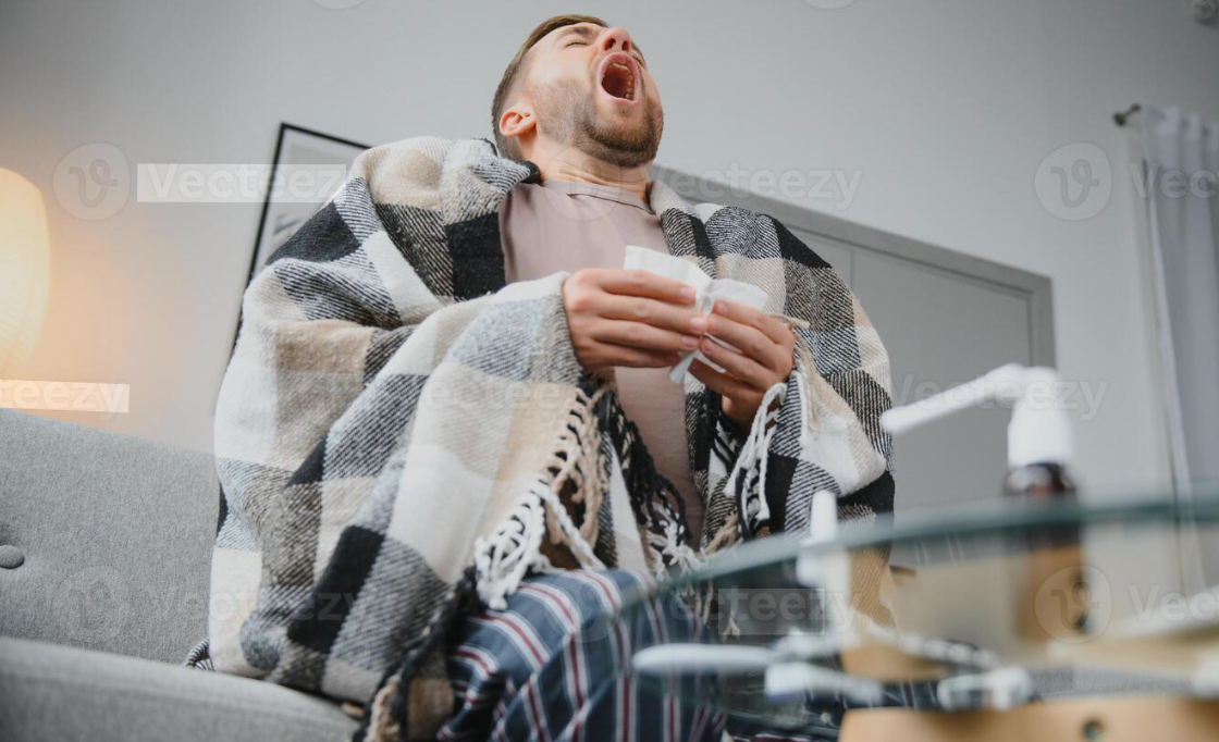 Homem espirrando enquanto segura um lenço, envolto em um cobertor no sofá, com sintomas de alergia