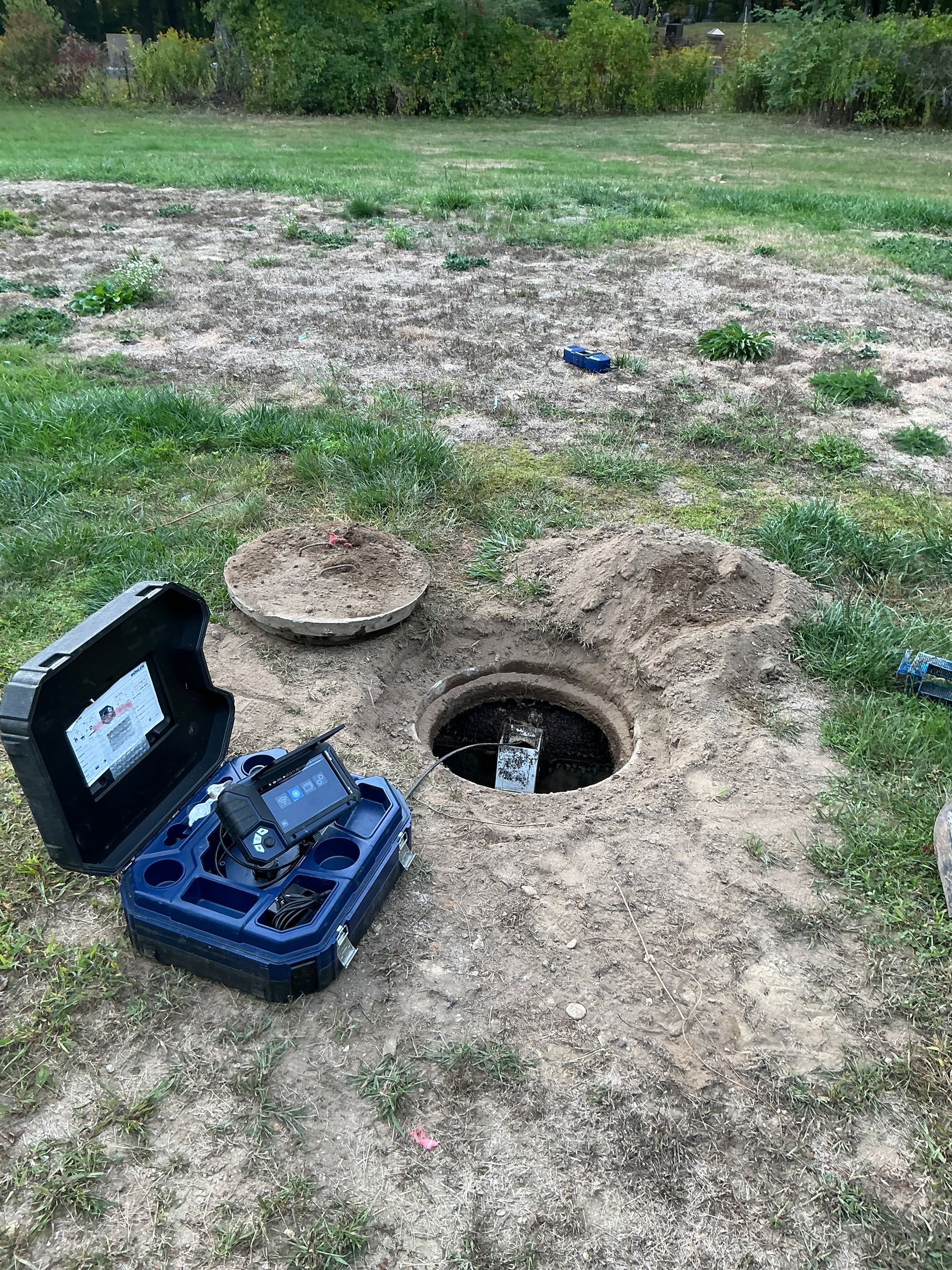 Open septic tank with equipment case nearby on grassy ground.