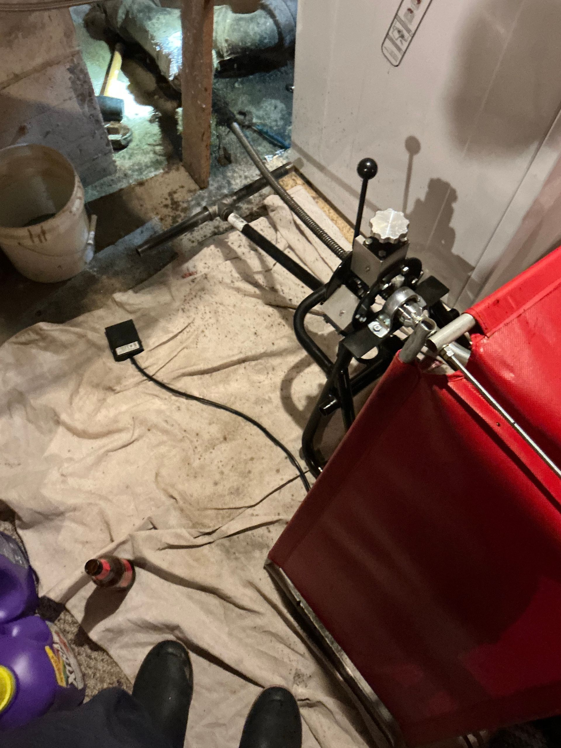 Red machine with metal pipes, tools, and a bucket inside a room with a white cloth on the floor.