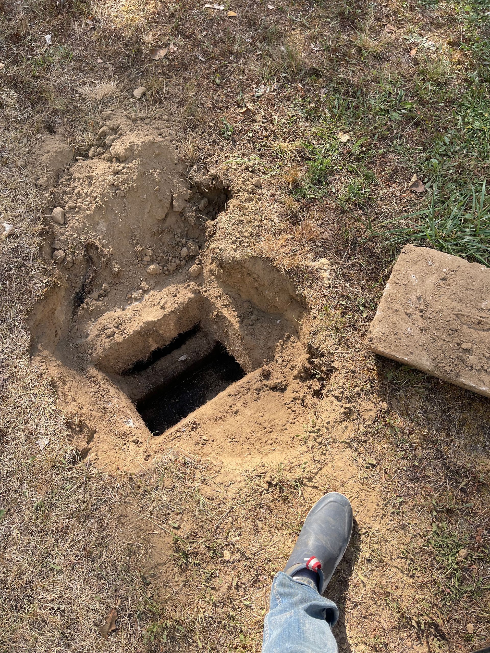 Hole in the ground with a square opening and a removed rectangular lid; a person's foot is visible.