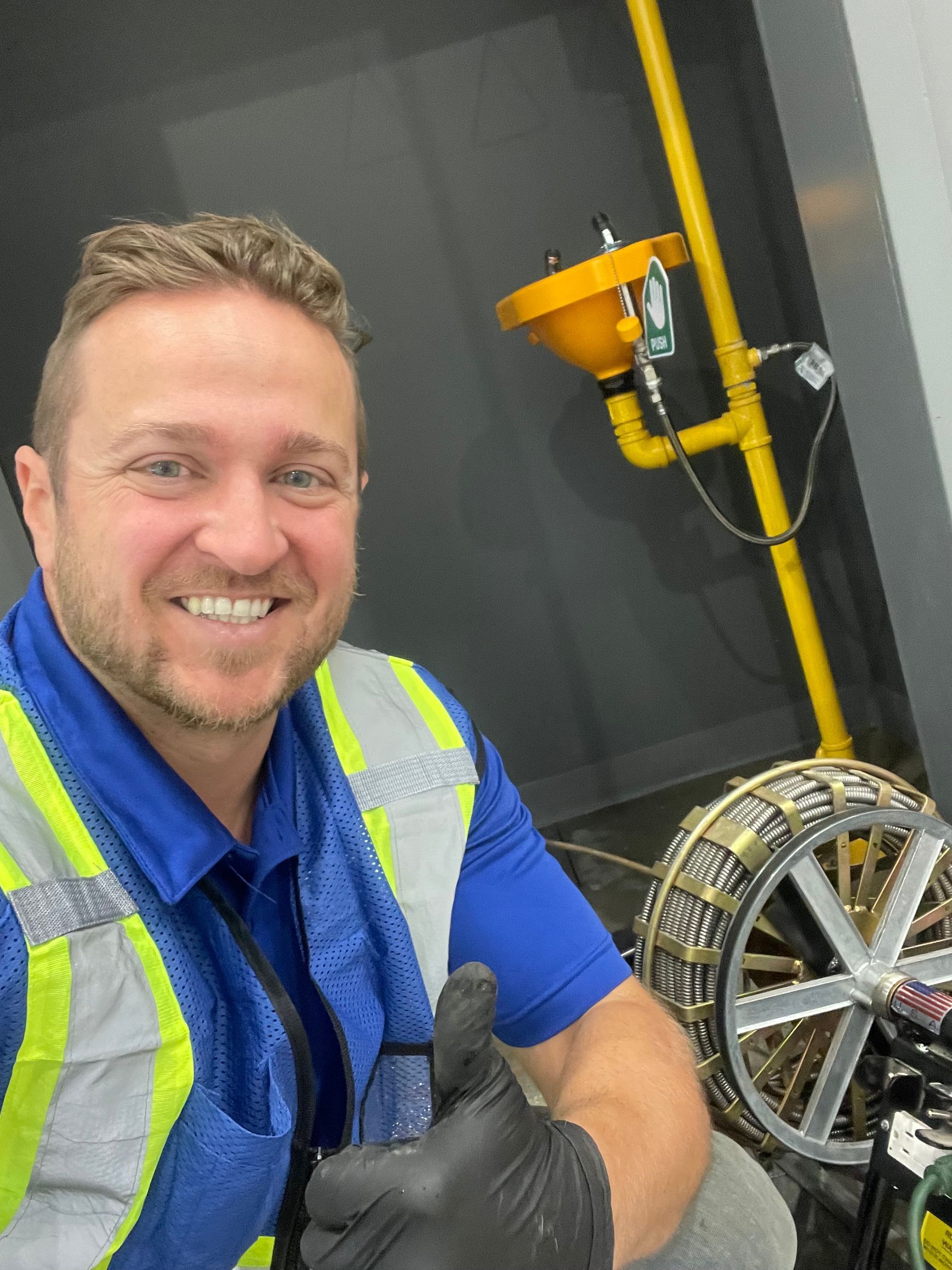 Man in work clothes gives thumbs up near yellow pipes and equipment.