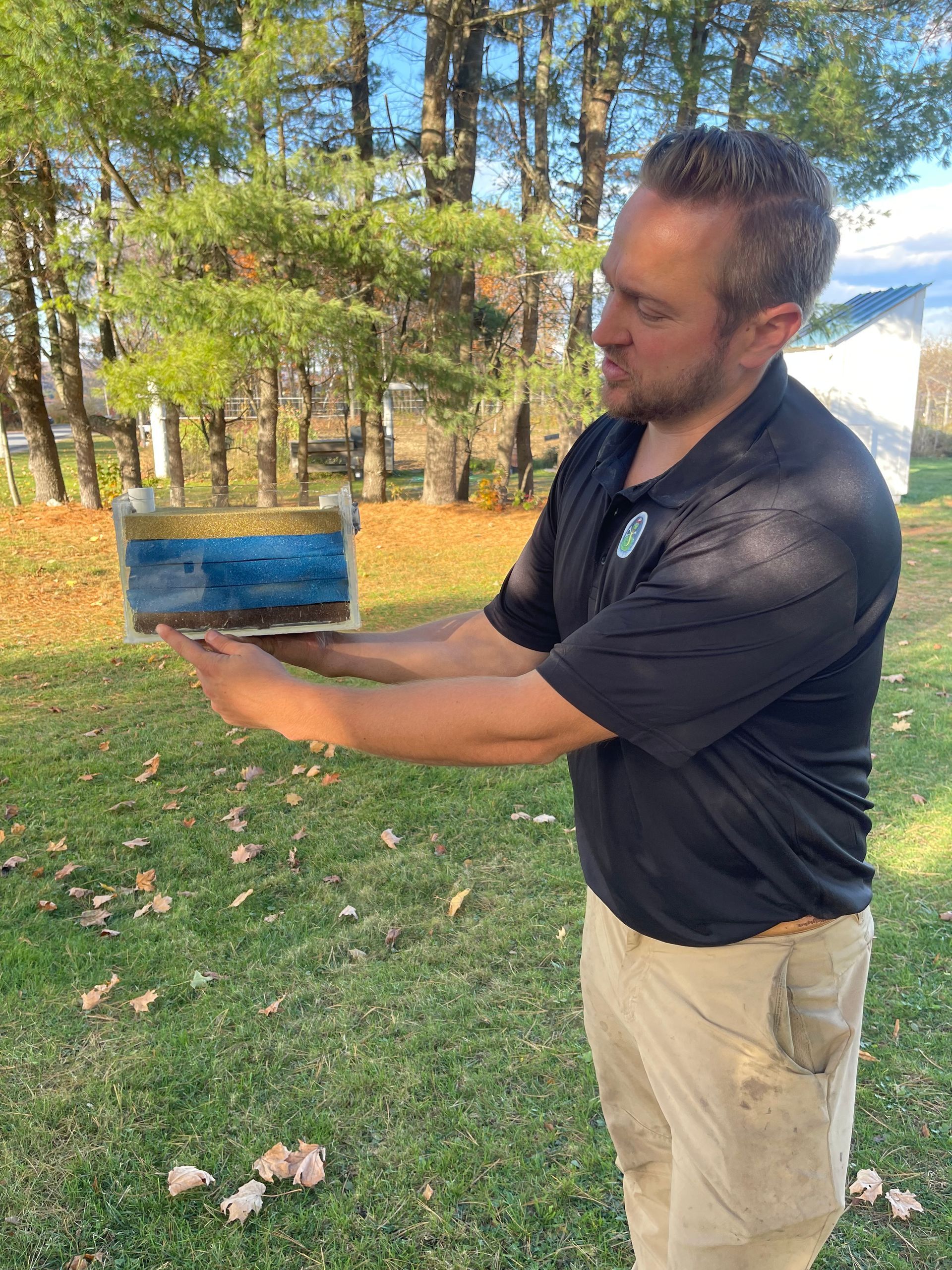 Man holding a rectangular container with blue water outdoors, grass, and trees in the background.