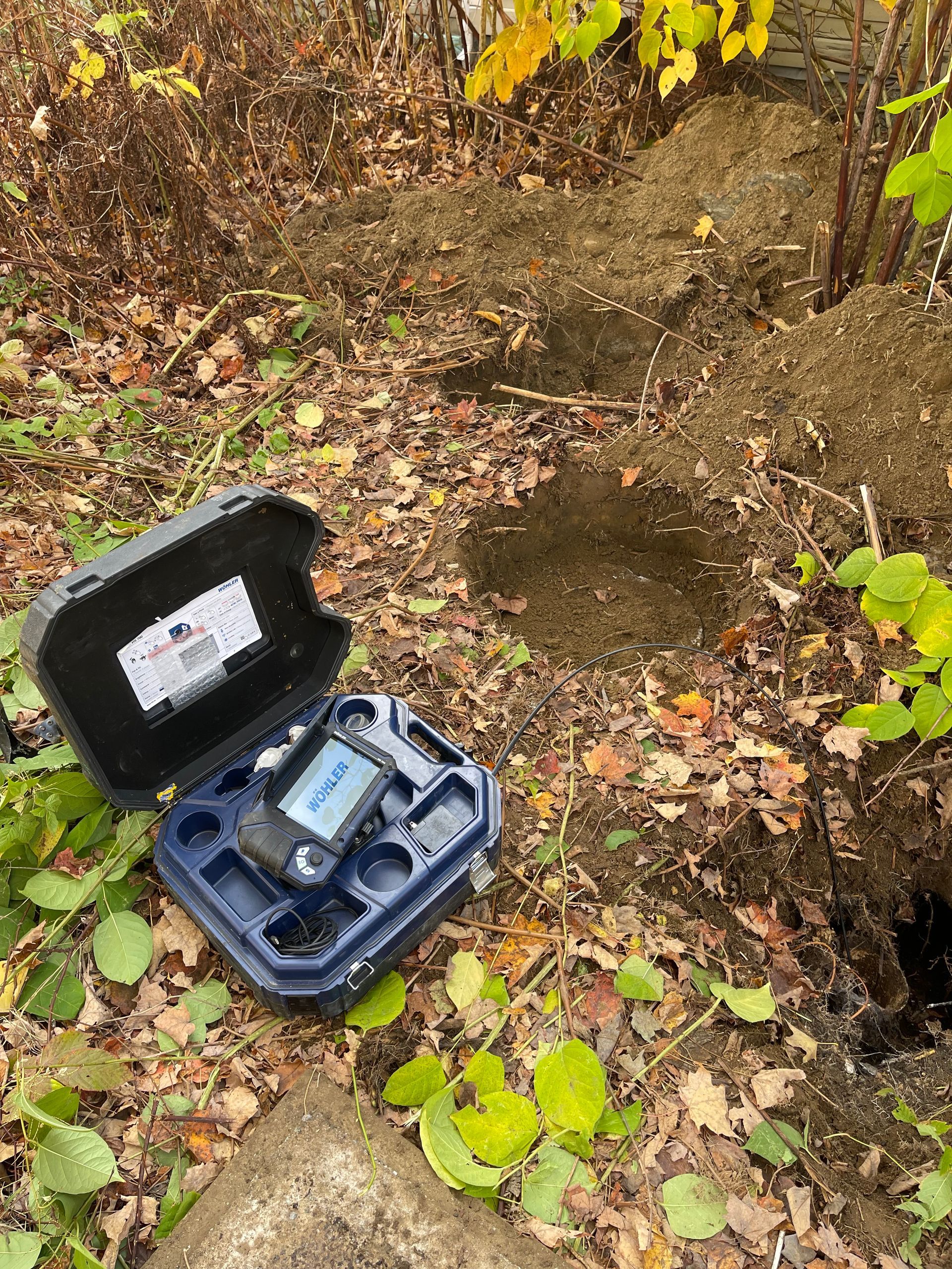 Open case with a device; ground with holes and soil pile; leafy foliage.
