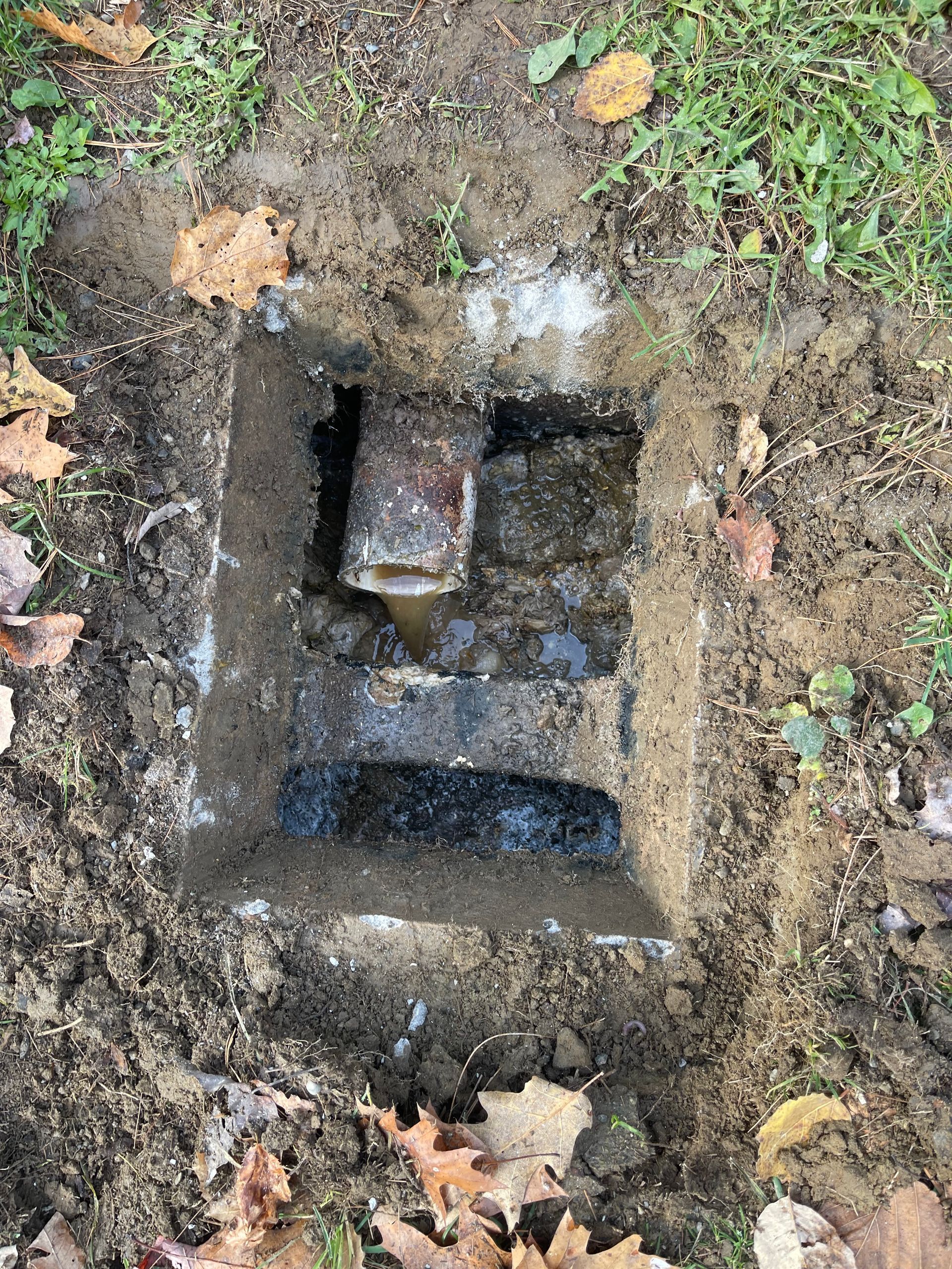 Open square hole in ground, revealing broken concrete and pipe. Surrounded by dirt and grass.