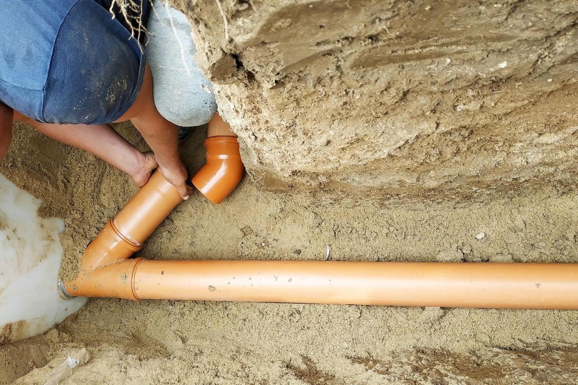 Person installing brown plastic drainage pipes in a trench filled with sand.