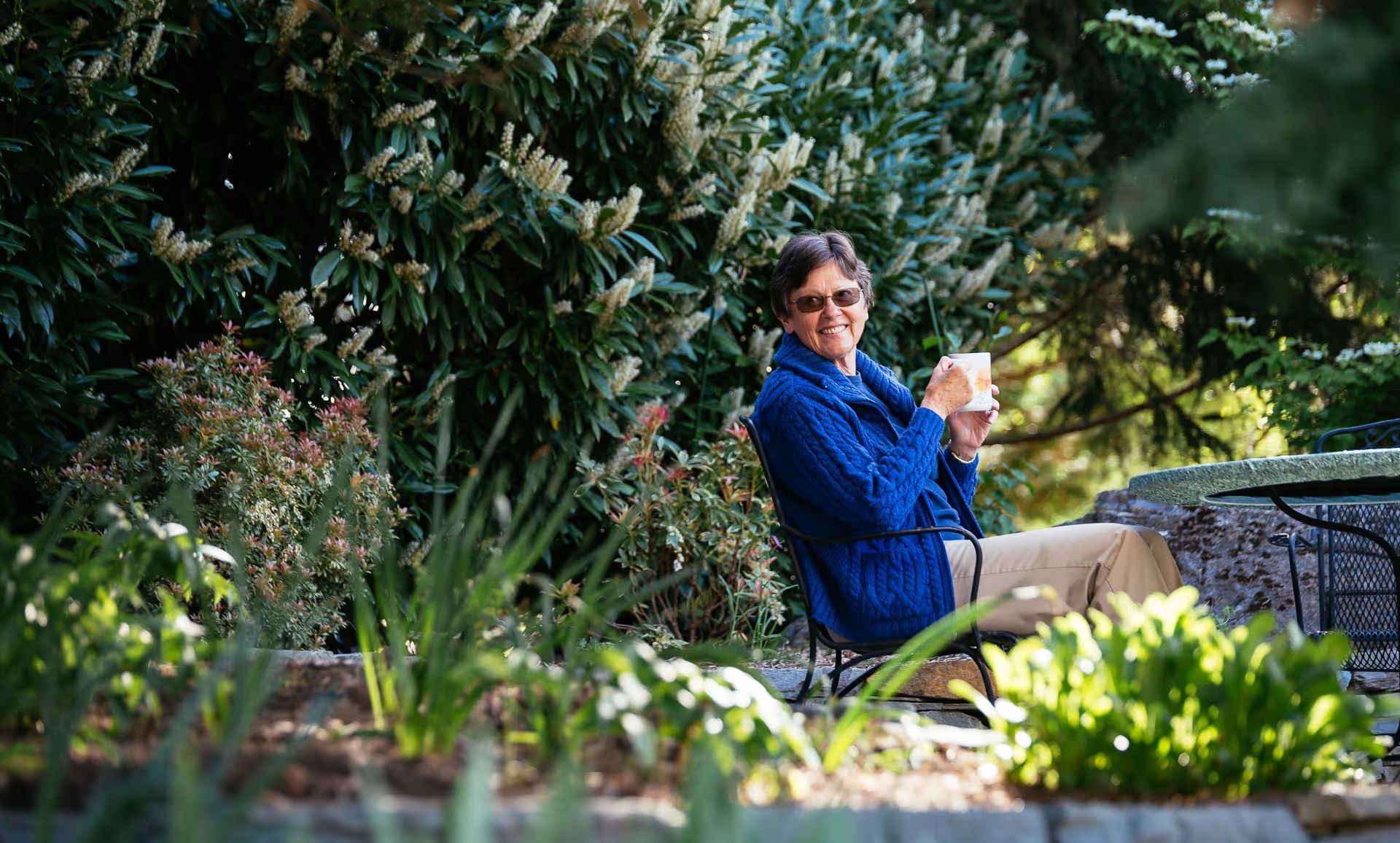 A woman is sitting on a bench in a garden holding a cup of coffee.