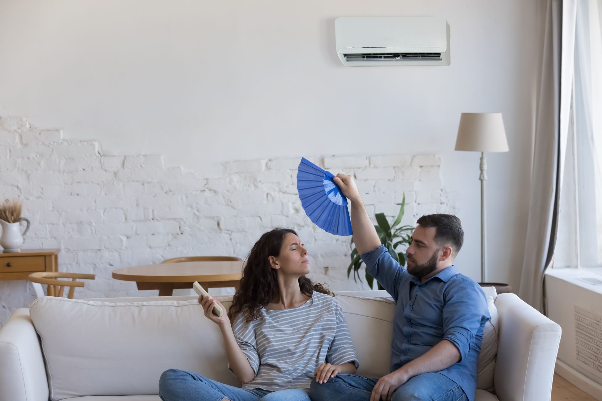 Couple fanning themselves on a couch under a wall-mounted AC unit.