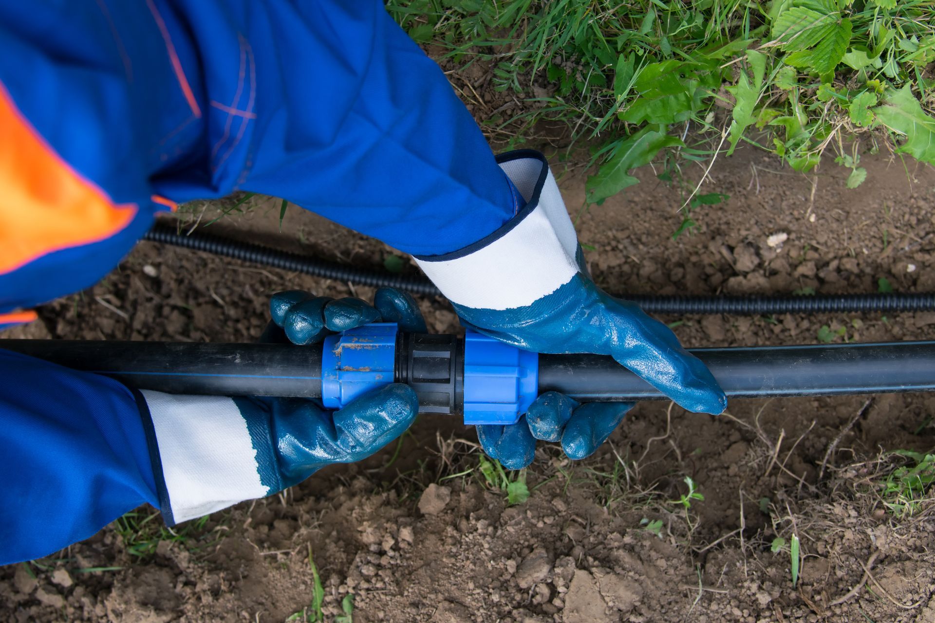 Person in blue work suit and gloves connecting black pipe with blue connector outdoors.