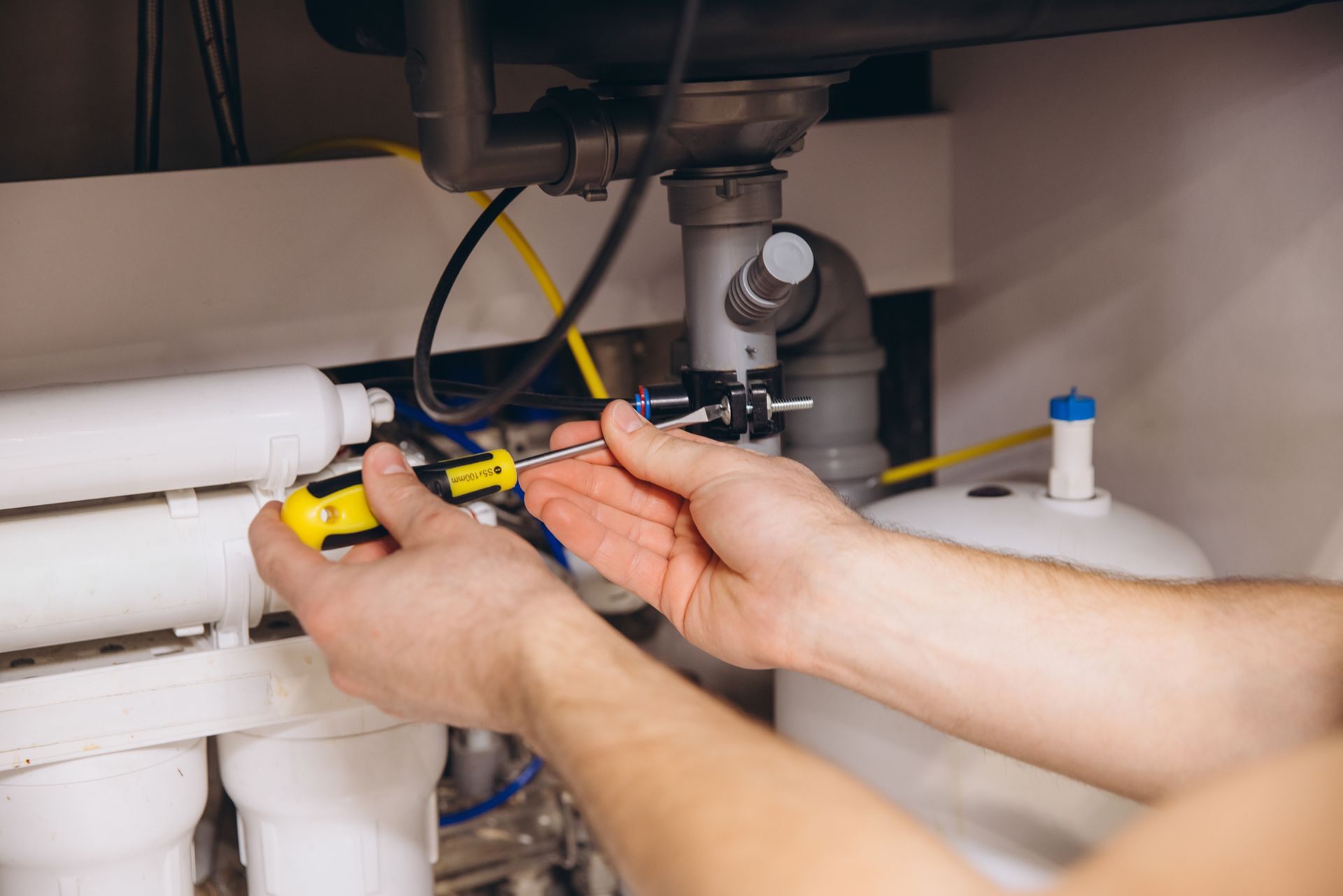 Hands using a screwdriver on plumbing under a sink with white pipes and a water filter.