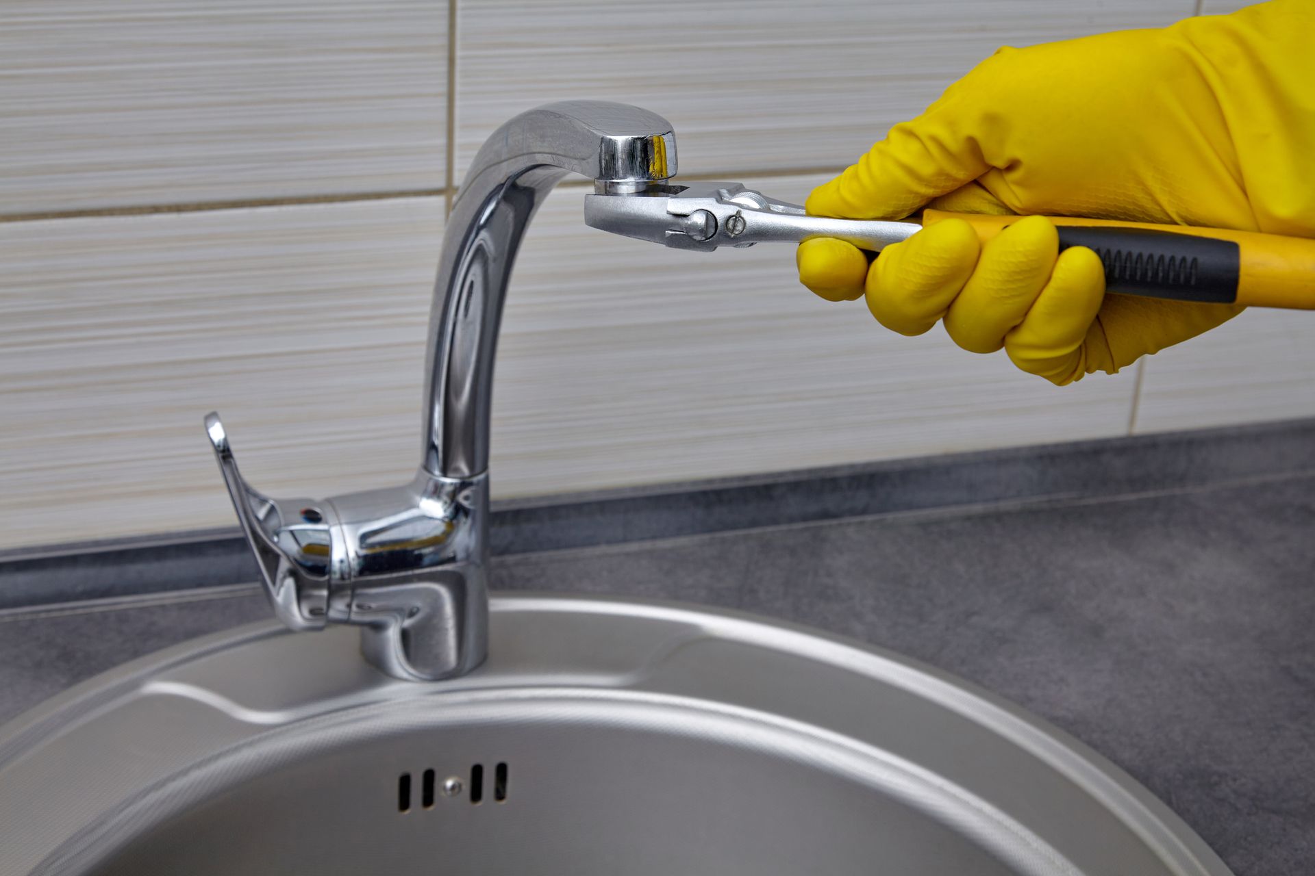 A person in yellow gloves uses a wrench to tighten a faucet in a stainless steel sink.