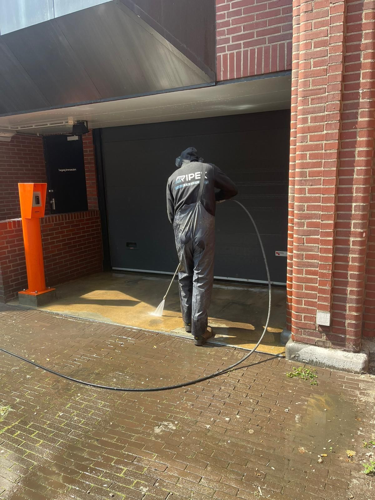 Een persoon in een donkere overall reinigt met een hogedrukreiniger een betonnen oppervlak in de buurt van een bakstenen gebouw met een garagedeur.