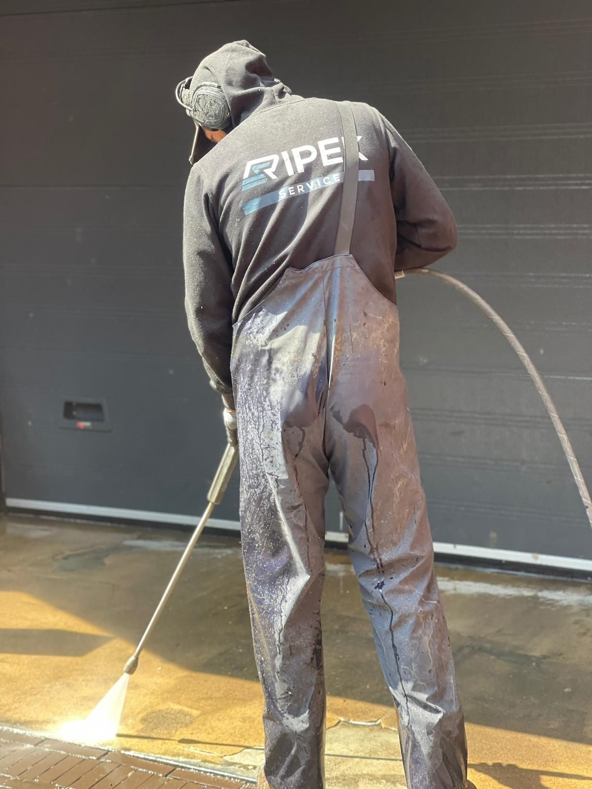 Een persoon in een zwarte waterdichte overall reinigt met een hogedrukreiniger een betonnen oppervlak naast een donkergrijze garagedeur.