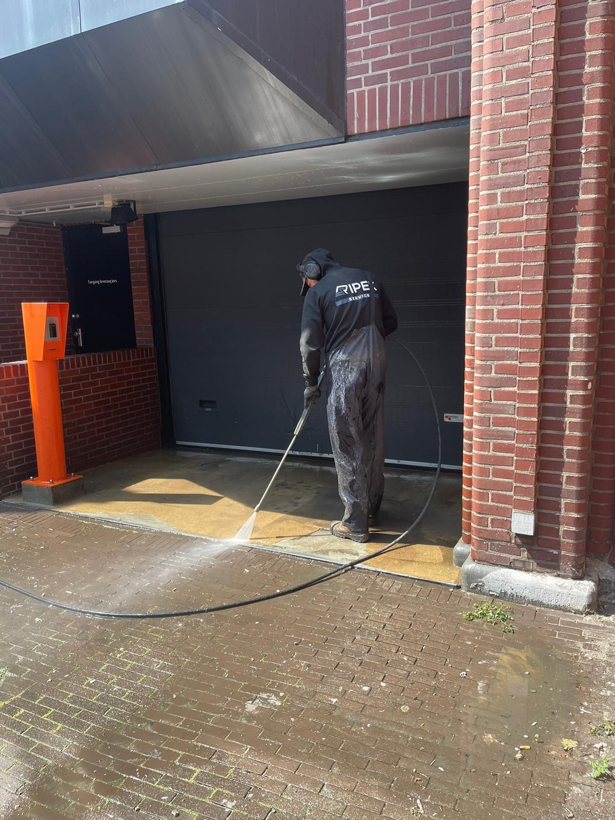 Persoon in overall reinigt met een hogedrukreiniger een betonnen oppervlak voor een grijze garagedeur. Bakstenen gebouw.