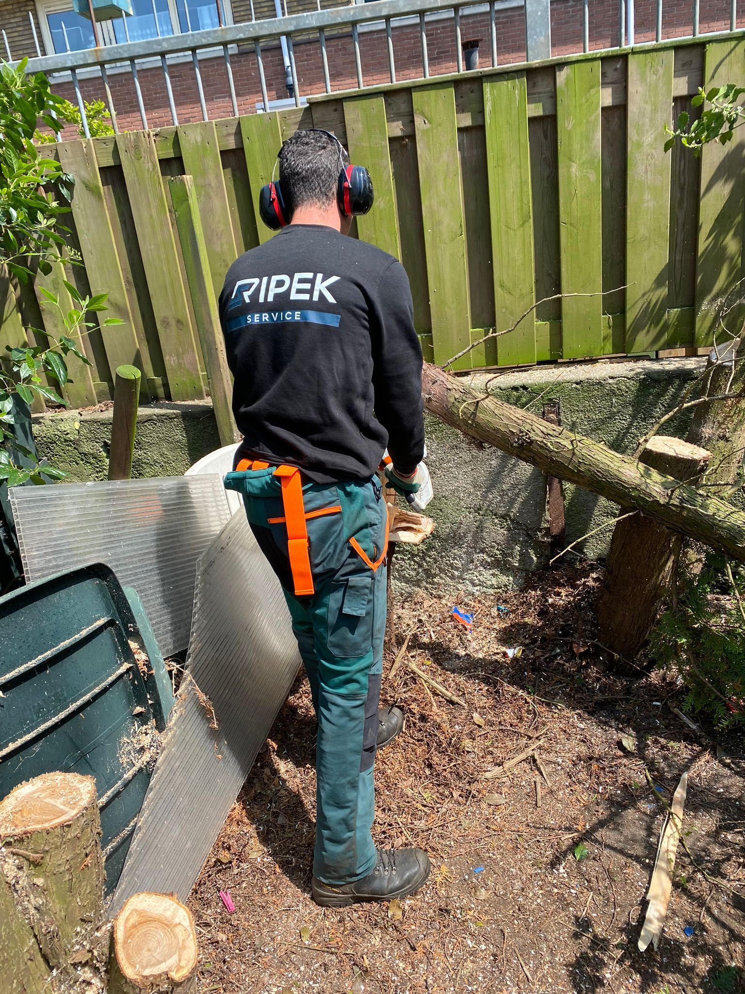 Een man zaagt met een kettingzaag een boomstam door in een achtertuin, terwijl hij veiligheidsuitrusting draagt.