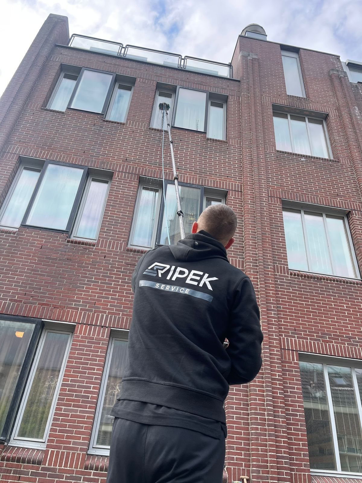 Een persoon maakt de ramen van een bakstenen gebouw schoon met een borstel met lange steel en wateraanvoer. De persoon draagt ​​een zwarte trui.