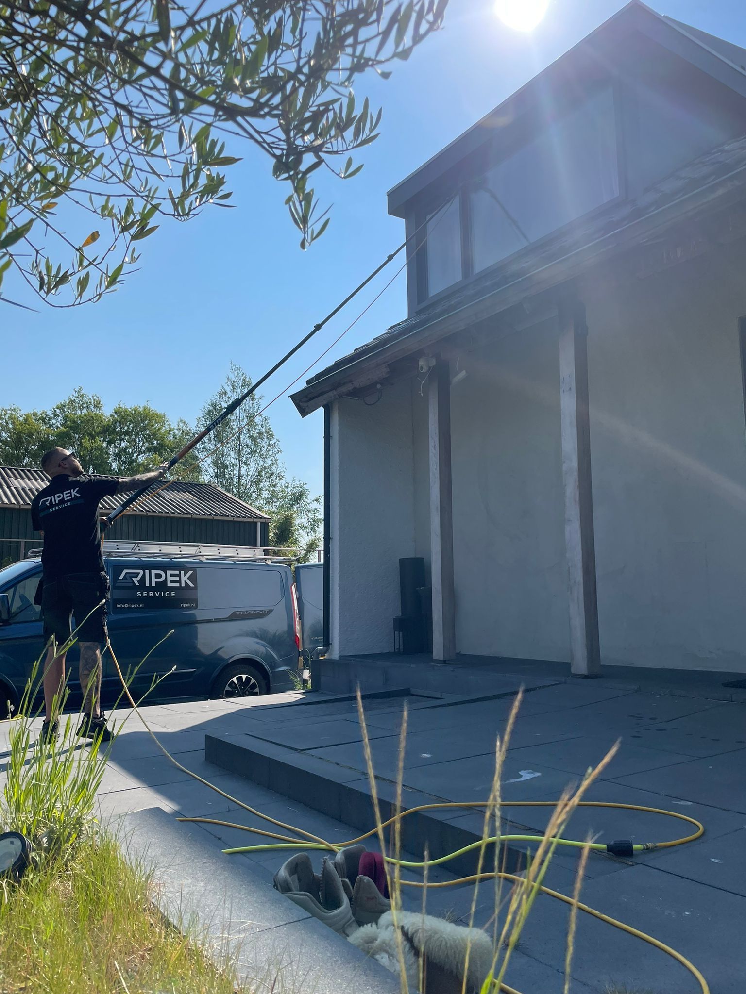 Een man maakt de ramen van een huis schoon met een gereedschap met een lange steel. Er staat een busje in de buurt geparkeerd. Zonnige buitenomgeving.
