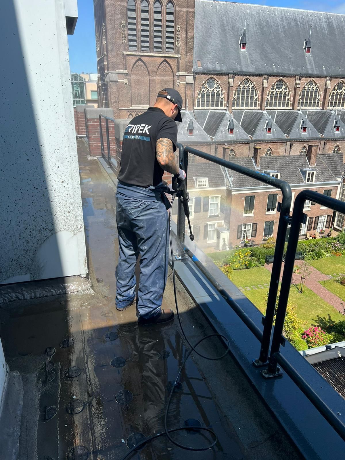 Een man reinigt met een hogedrukreiniger een dak, met op de achtergrond een gebouw en een kerk.