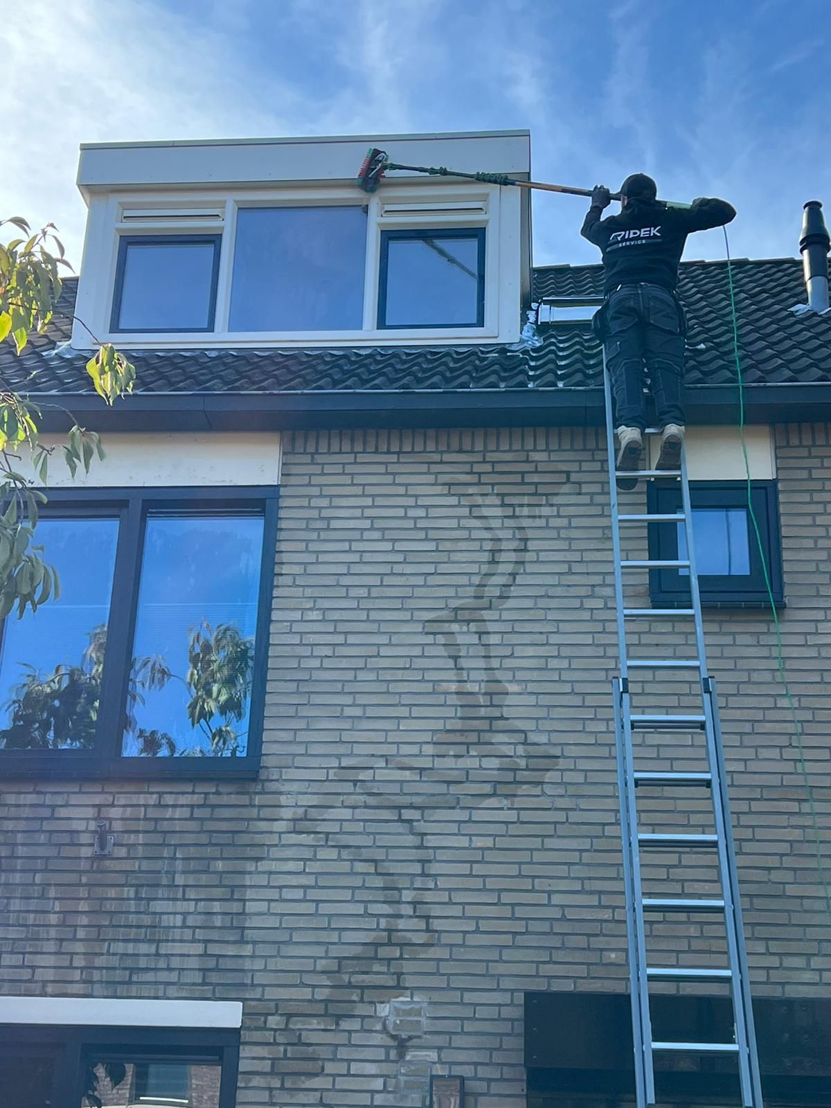 Een persoon maakt ramen schoon vanaf een ladder, met behulp van een borstel met lange steel, op een bakstenen gebouw met donker omlijste ramen.