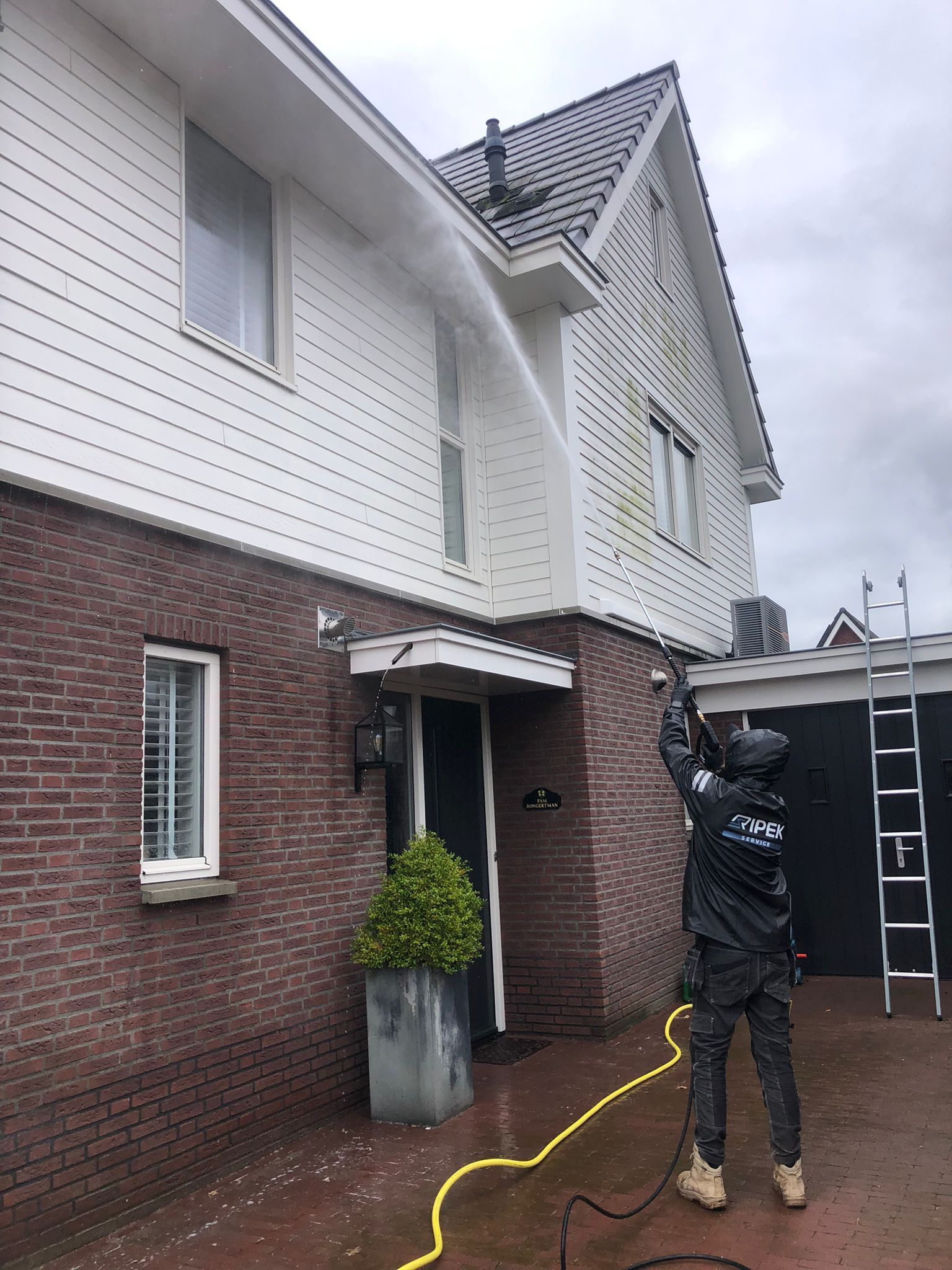 Een man reinigt met een hogedrukreiniger een twee verdiepingen tellend huis met witte gevelbekleding en een bakstenen fundering; het is een bewolkte dag.