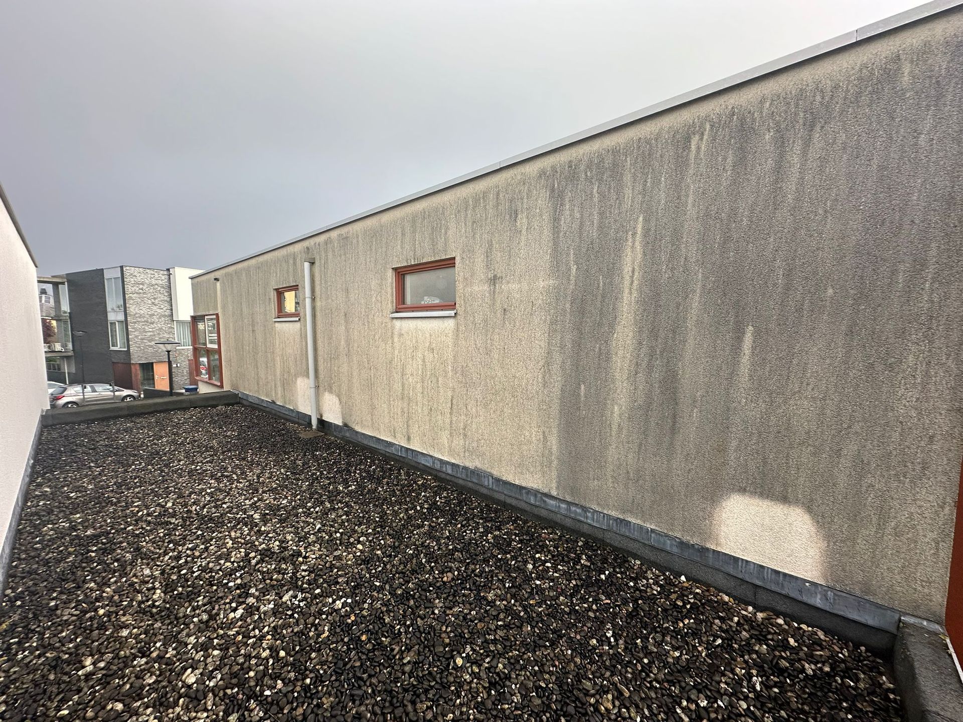 Een grindpad naast een lang, lichtgrijs gebouw met twee kleine ramen en een metalen afwerking.