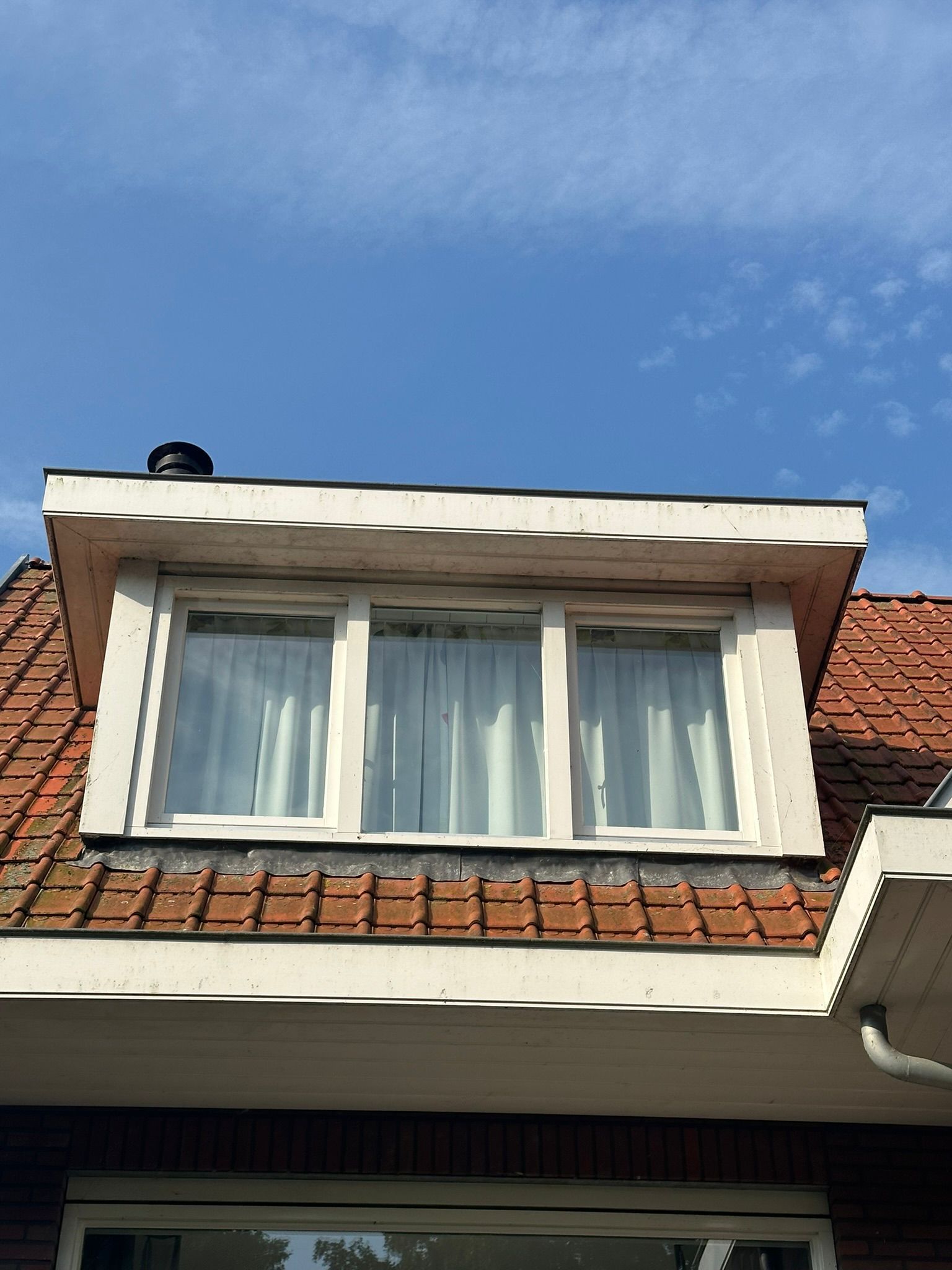 Dakkapel met drie ruiten, witte omlijsting, rood pannendak, tegen een blauwe lucht.
