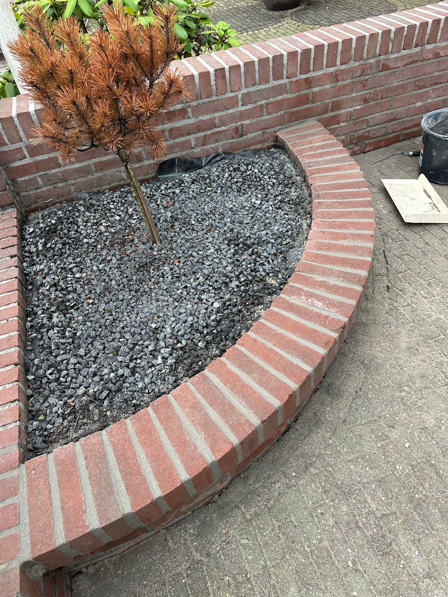 Bakstenen plantenbak met gebogen rand, gevuld met grijs grind, en een klein dood boompje.