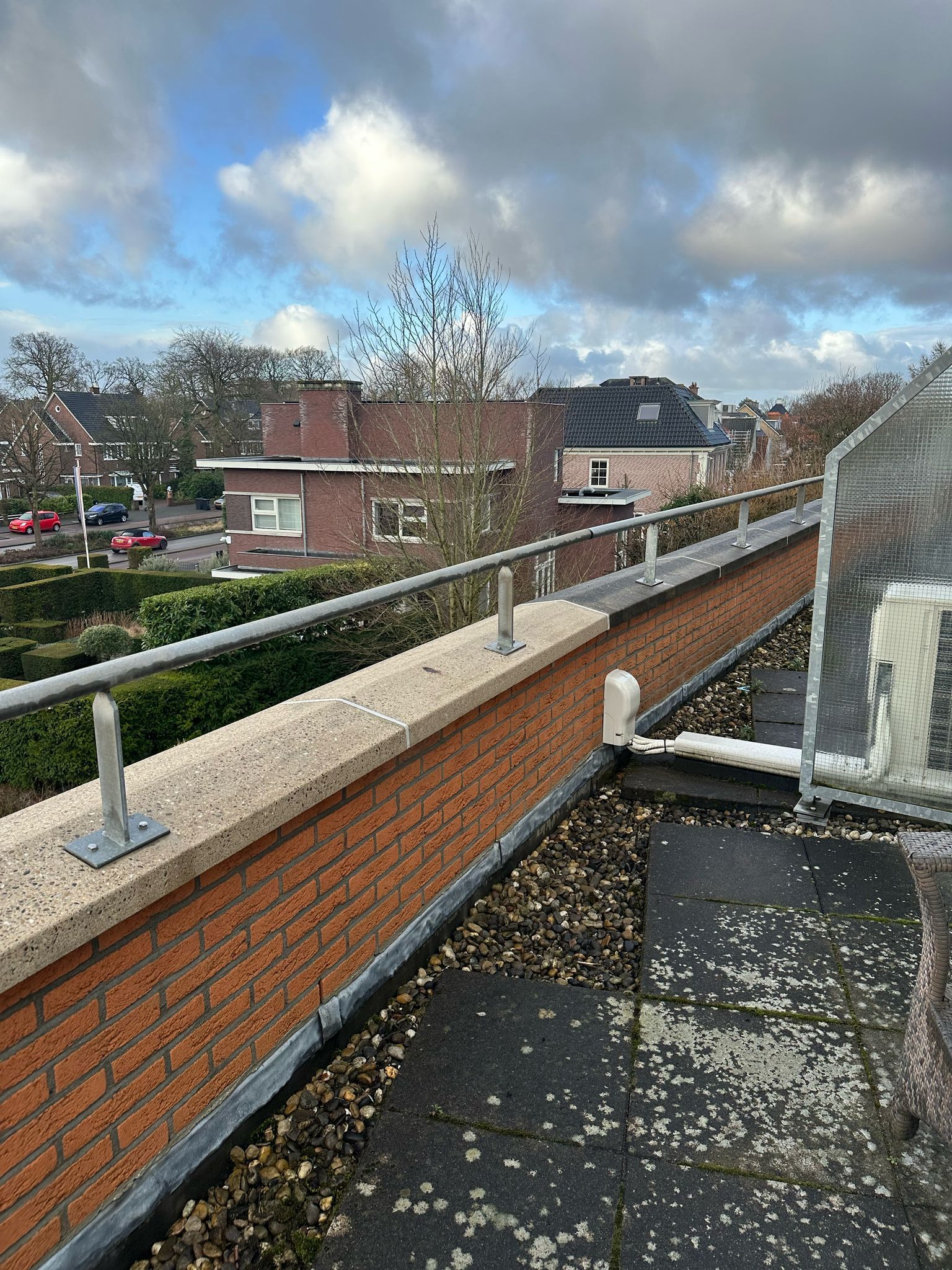 Een bakstenen dak met een balustrade, een blauwe lucht en gebouwen in de verte.