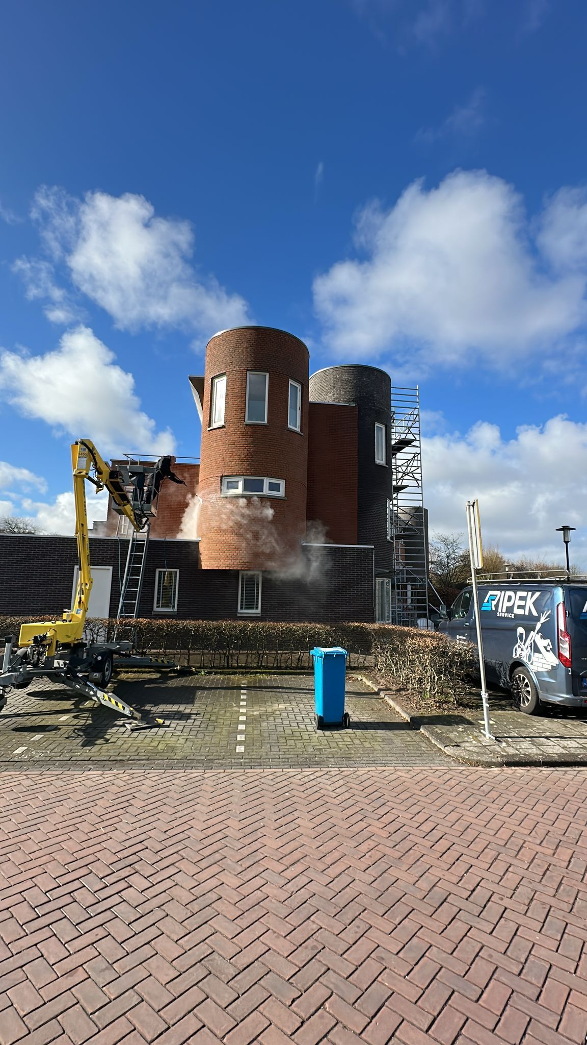 De sloop van een bakstenen gebouw met cilindrische torens; een graafmachine is aan het werk onder een blauwe hemel.