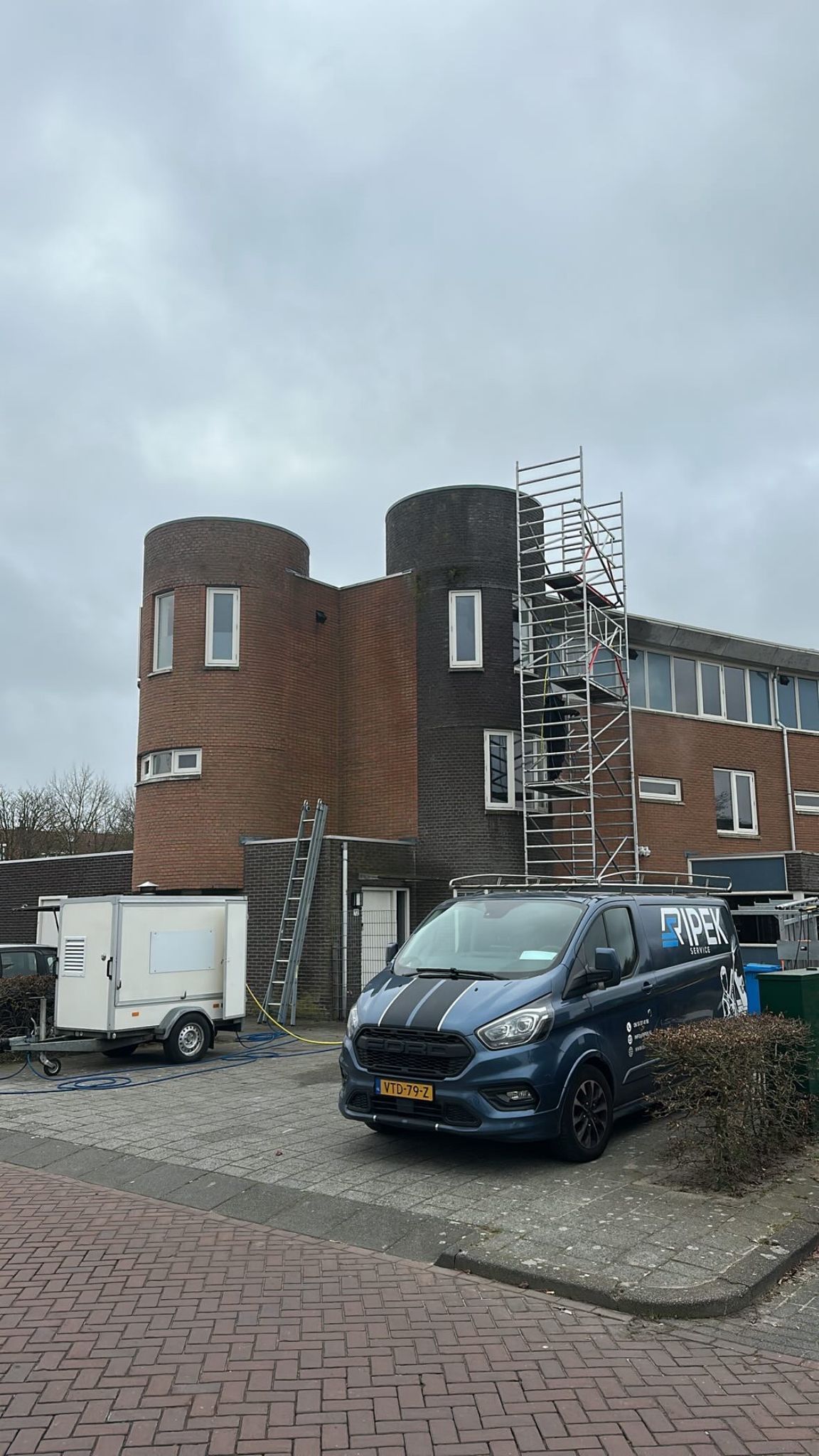 Donkerblauwe bestelwagen geparkeerd voor een bakstenen gebouw met steigers. Aanhangwagen links. Bewolkte lucht.