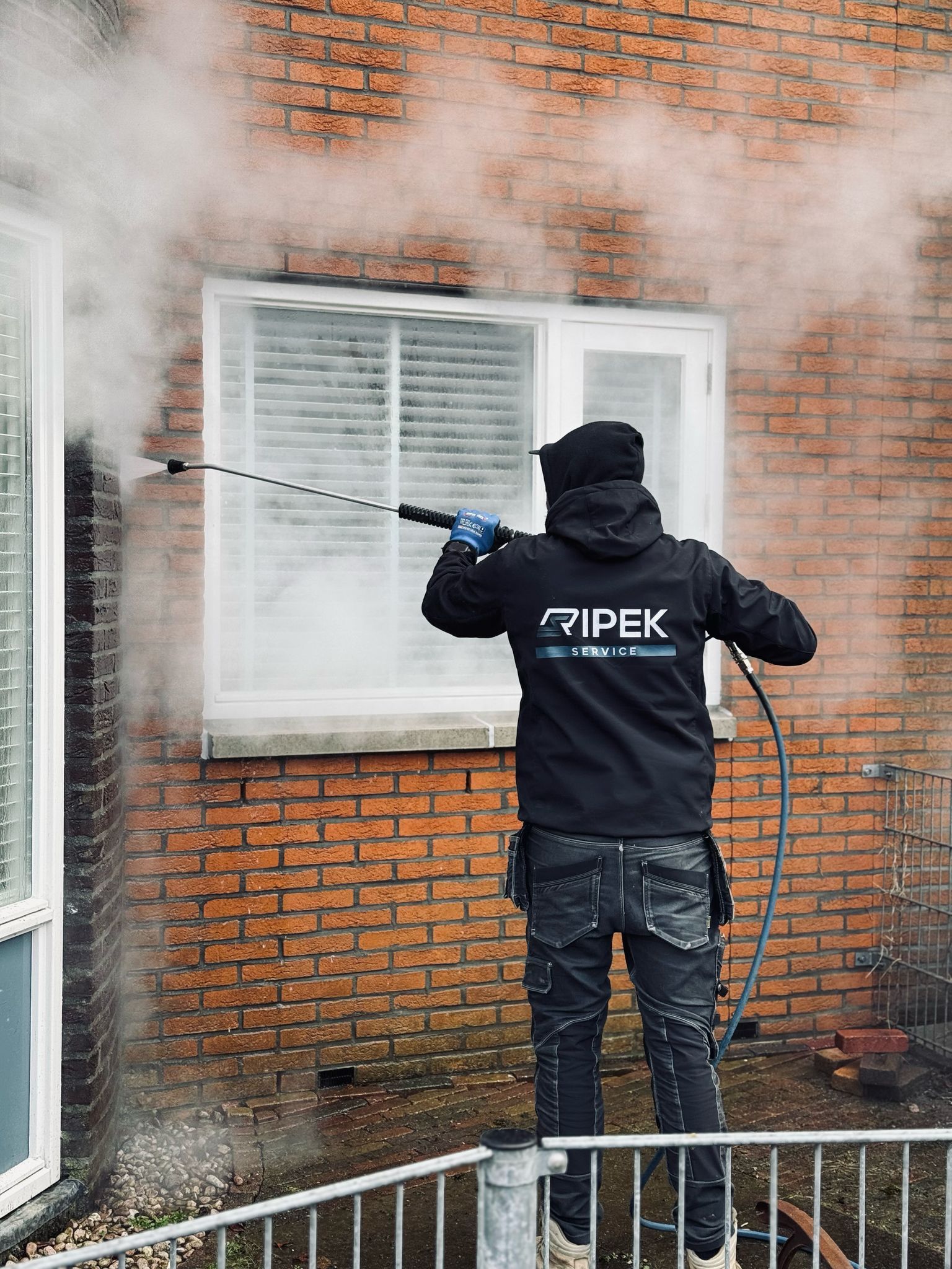 Iemand reinigt een raam van een bakstenen gebouw met een hogedrukreiniger, waardoor stoom ontstaat.
