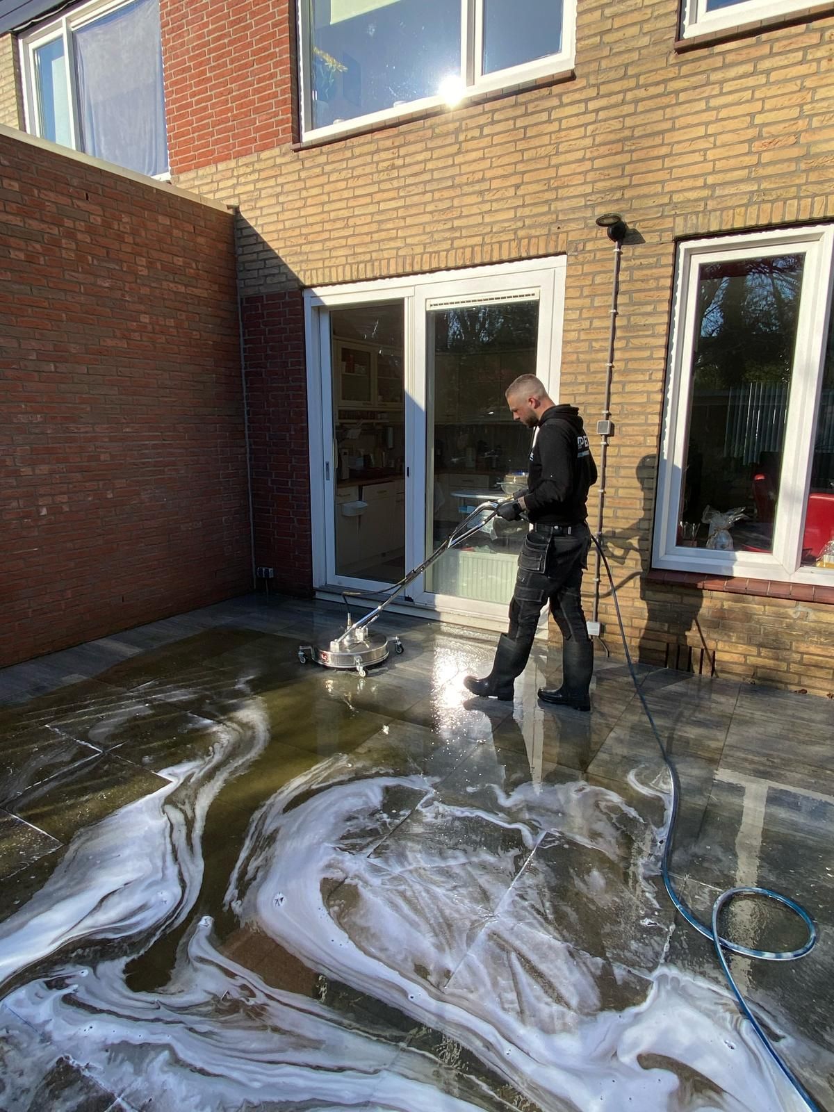 Iemand reinigt een terras met een hogedrukreiniger, waardoor er zeepsop op het oppervlak ontstaat, vlakbij een bakstenen gebouw.