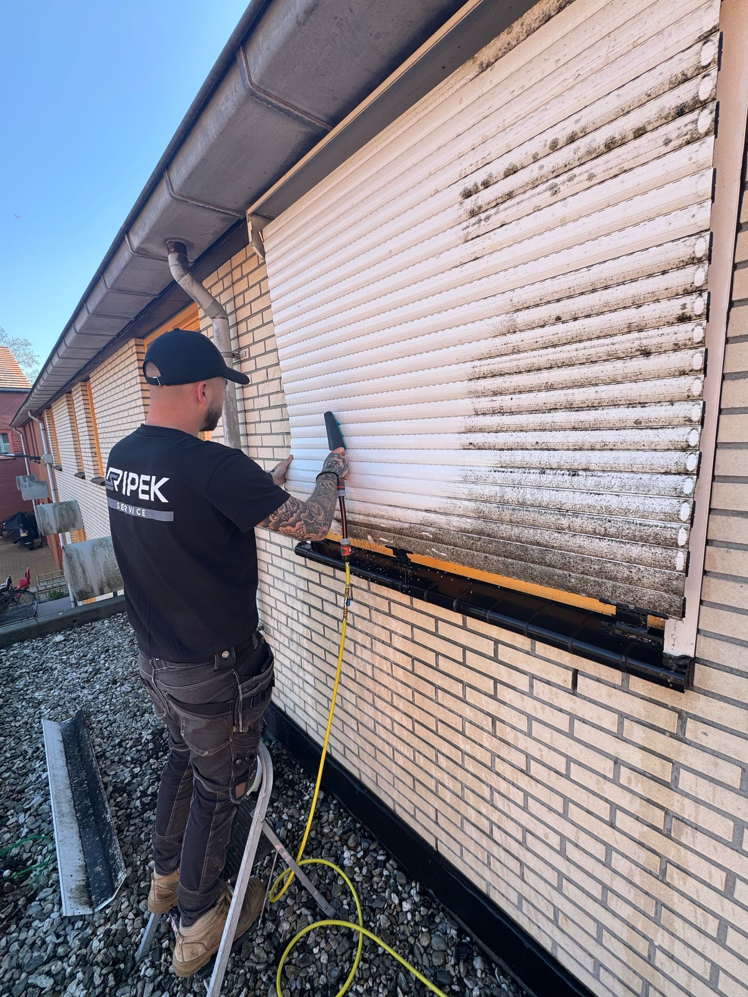 Een man reinigt met een hogedrukreiniger een vuil wit rolluik aan de buitenkant van een bakstenen gebouw.