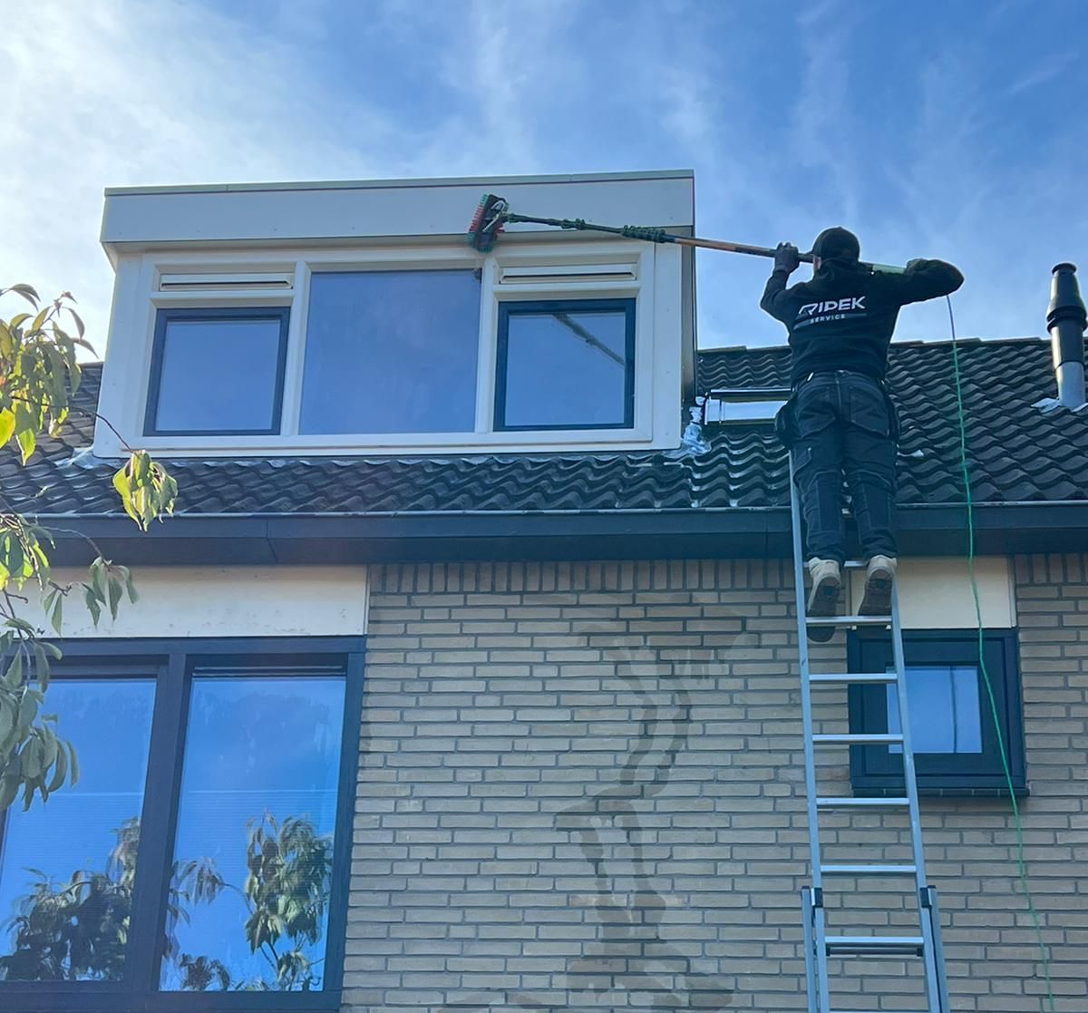 Een persoon maakt vanaf een ladder de ramen van een bakstenen huis schoon onder een blauwe hemel.