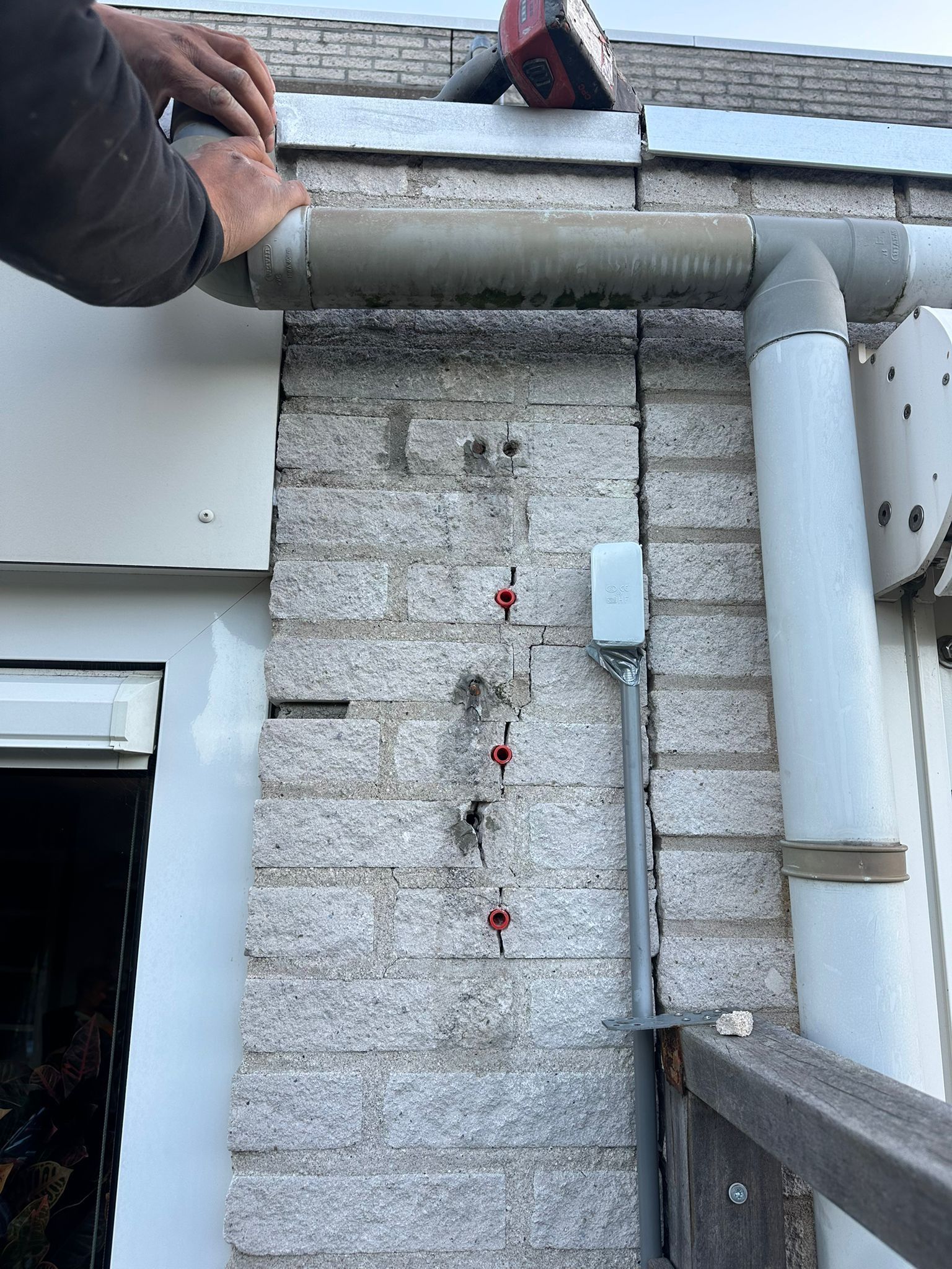 Man installeert een dakgoot op een bakstenen muur, rode laserwaterpas bovenaan. Verticale buis rechts.