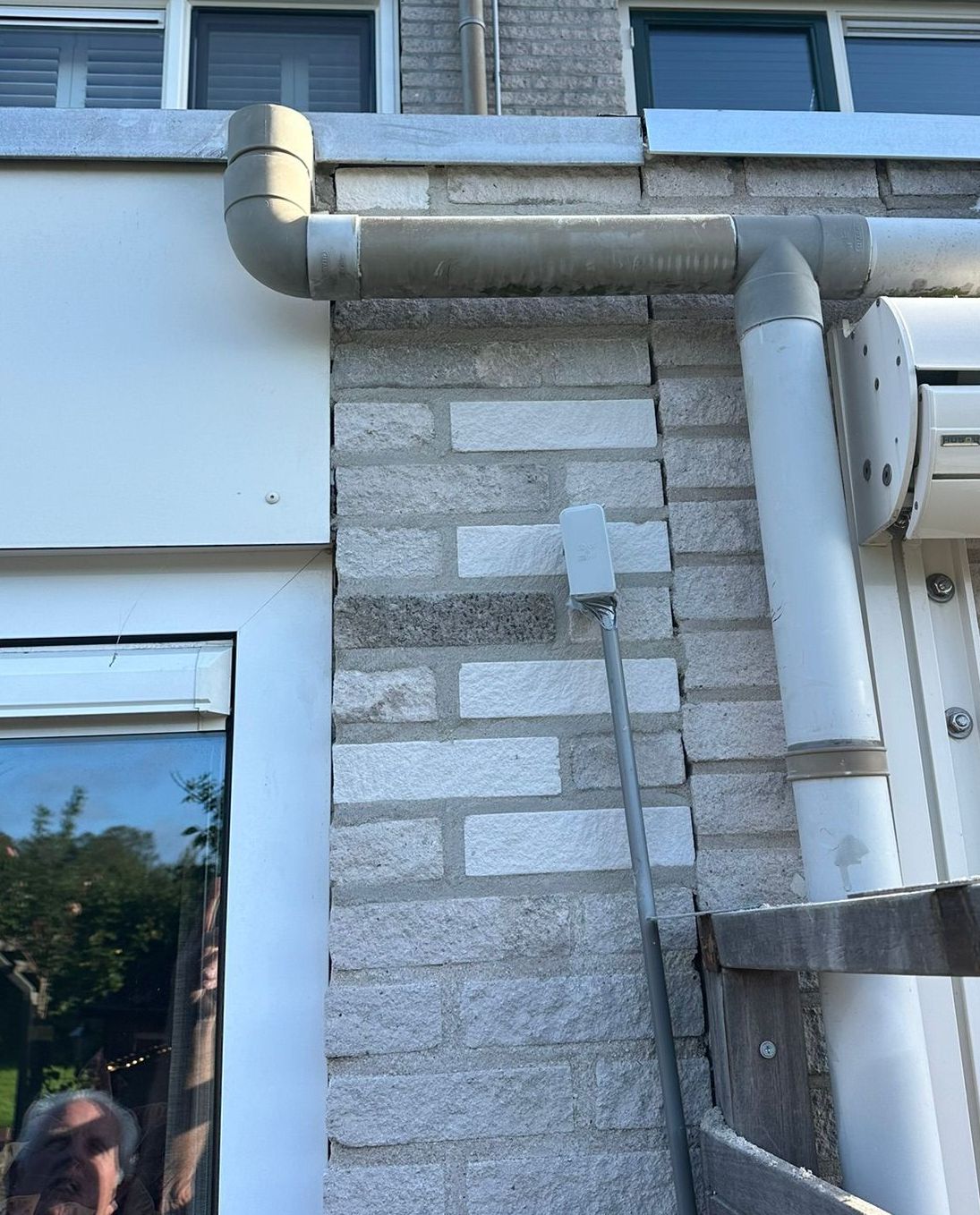 Bakstenen muur met witte leidingen en elektriciteitskast tegen het gebouw, raam zichtbaar.