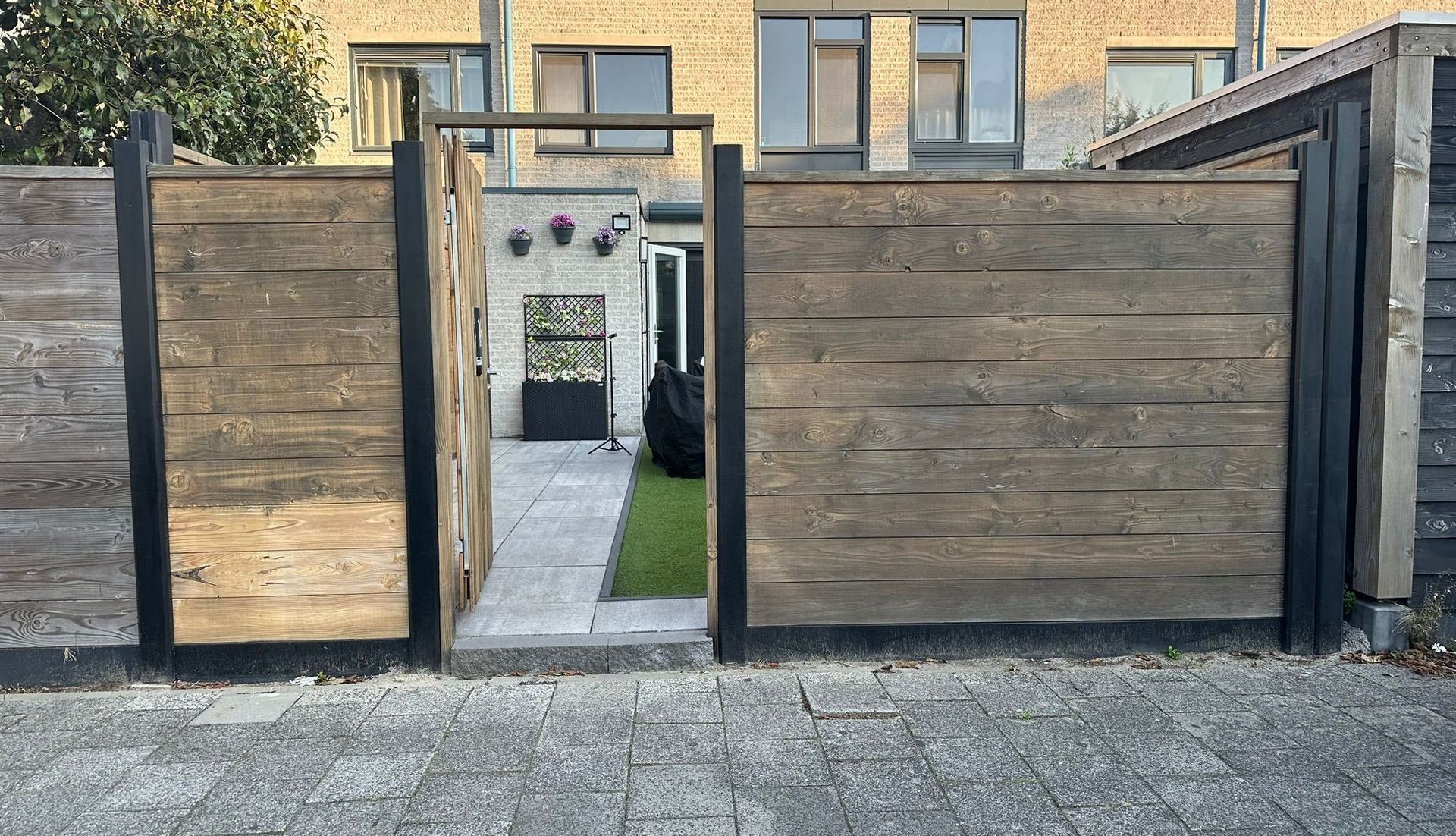 Houten schutting met open poort die toegang geeft tot een kleine binnenplaats met groen.