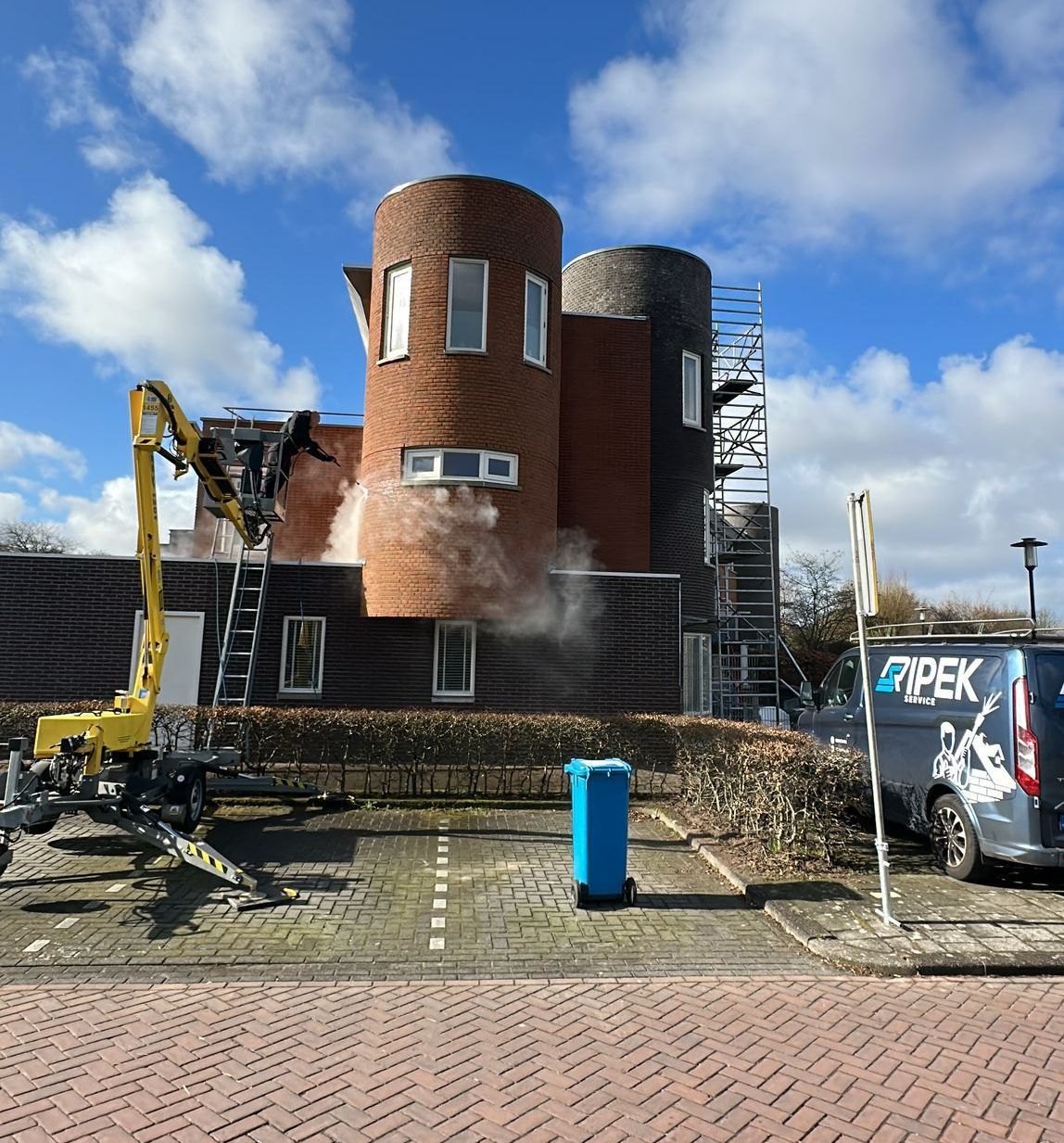 Een sloopgraafmachine sproeit water op een bakstenen gebouw met twee torentjes, op de achtergrond een blauwe hemel.