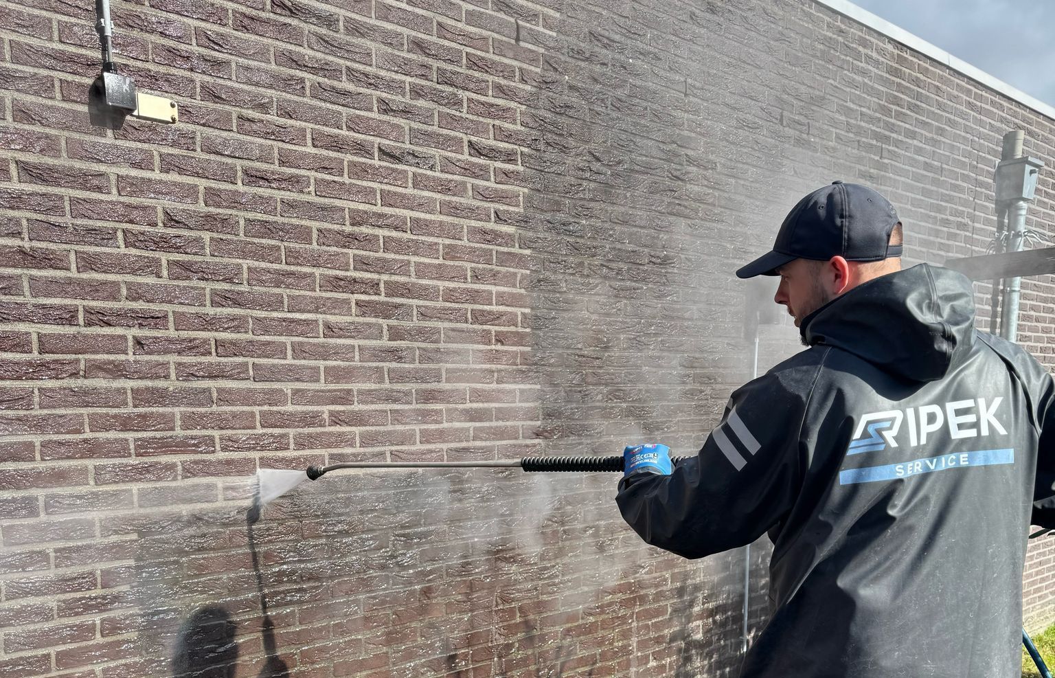 Man in regenkleding reinigt een bakstenen muur met een hogedrukreiniger.