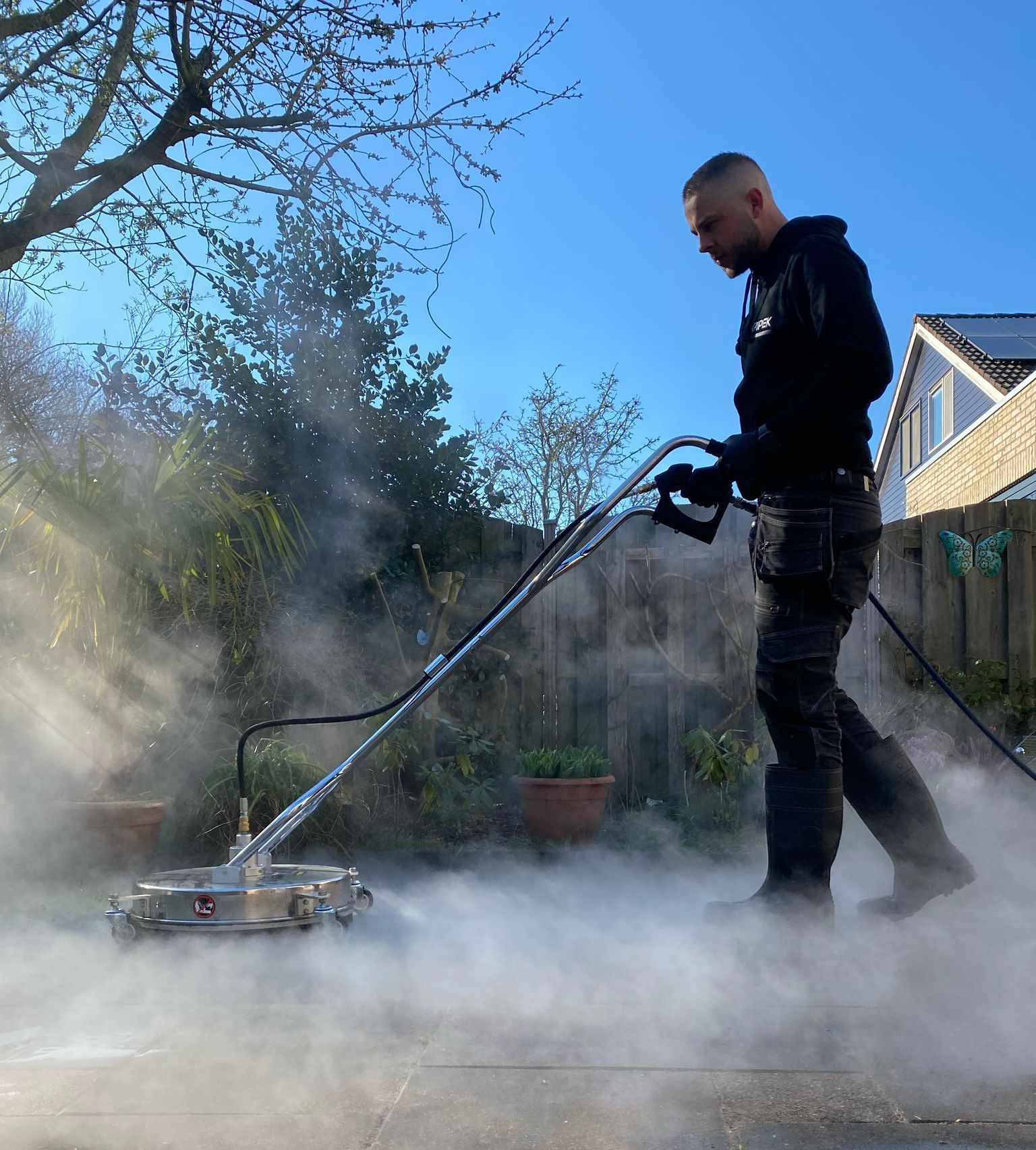 Een man reinigt een betonnen terras met een hogedrukreiniger, waardoor stoom ontstaat.