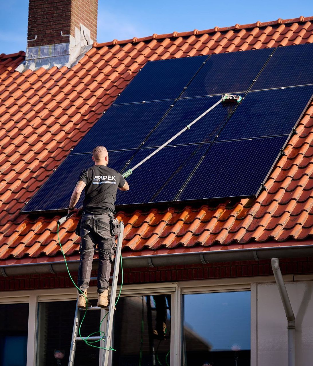 Een persoon die zonnepanelen op een rood pannendak schoonmaakt met een borstel met lange steel, staand op een ladder.