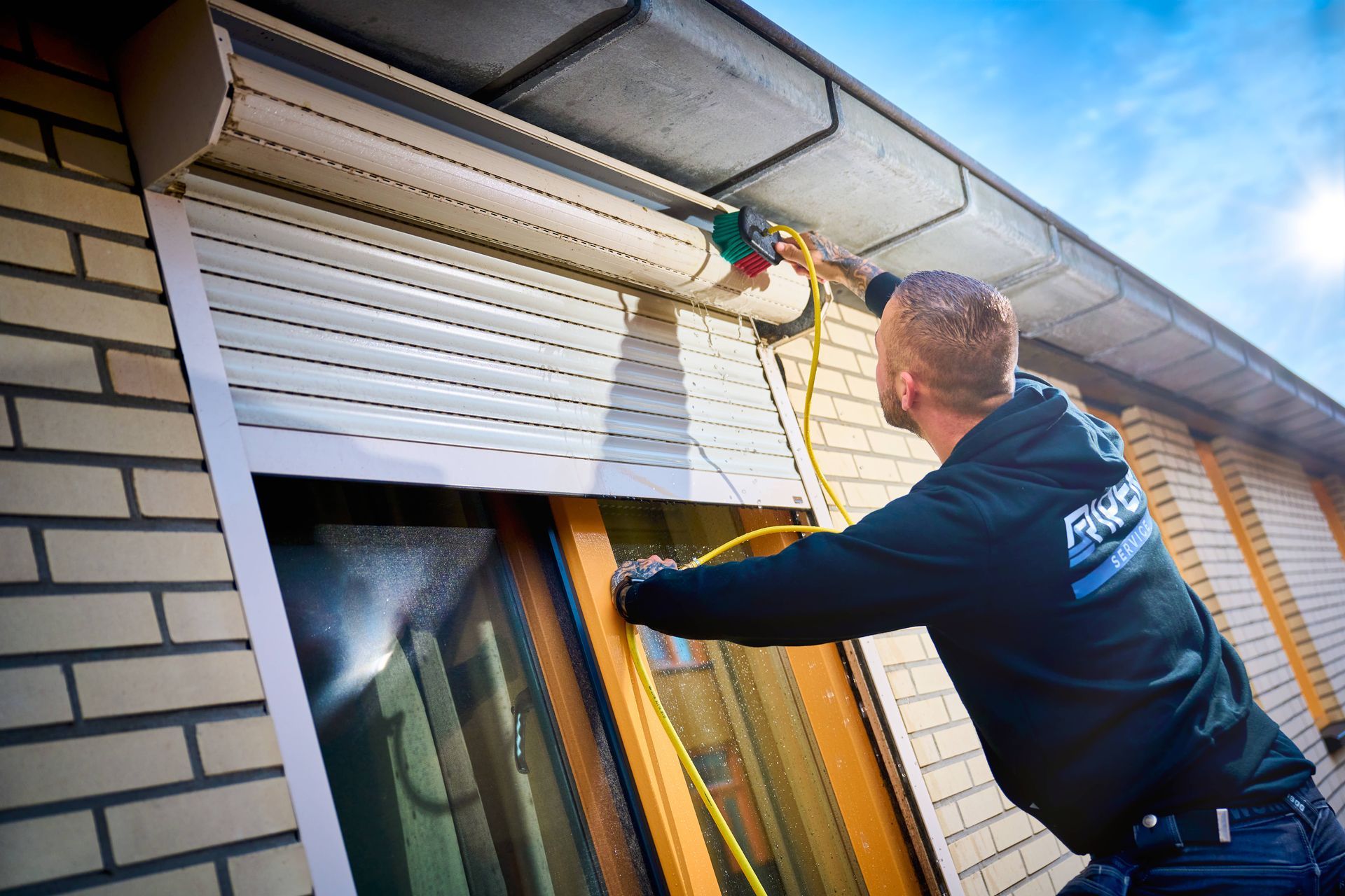 Een man is bezig met het repareren van een buitenluik. Hij houdt een elektrisch gereedschap vast; het raam is zichtbaar.
