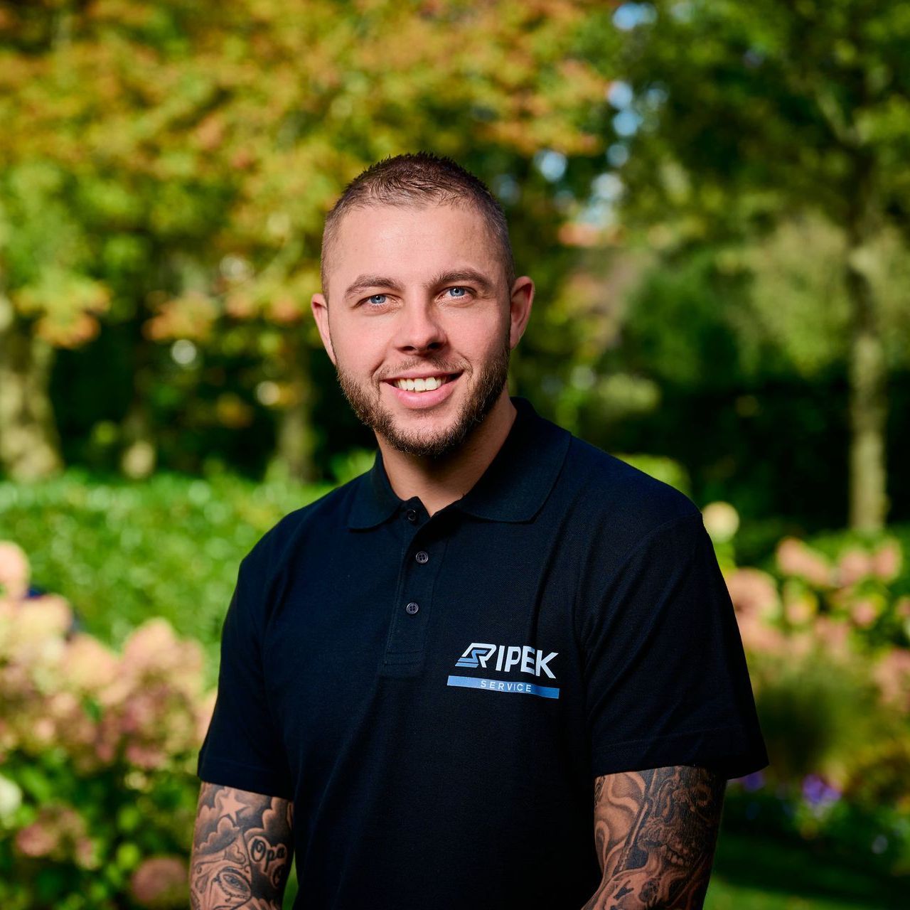 Man met tatoeages in een zwart poloshirt, lachend buiten voor een bladerdak.