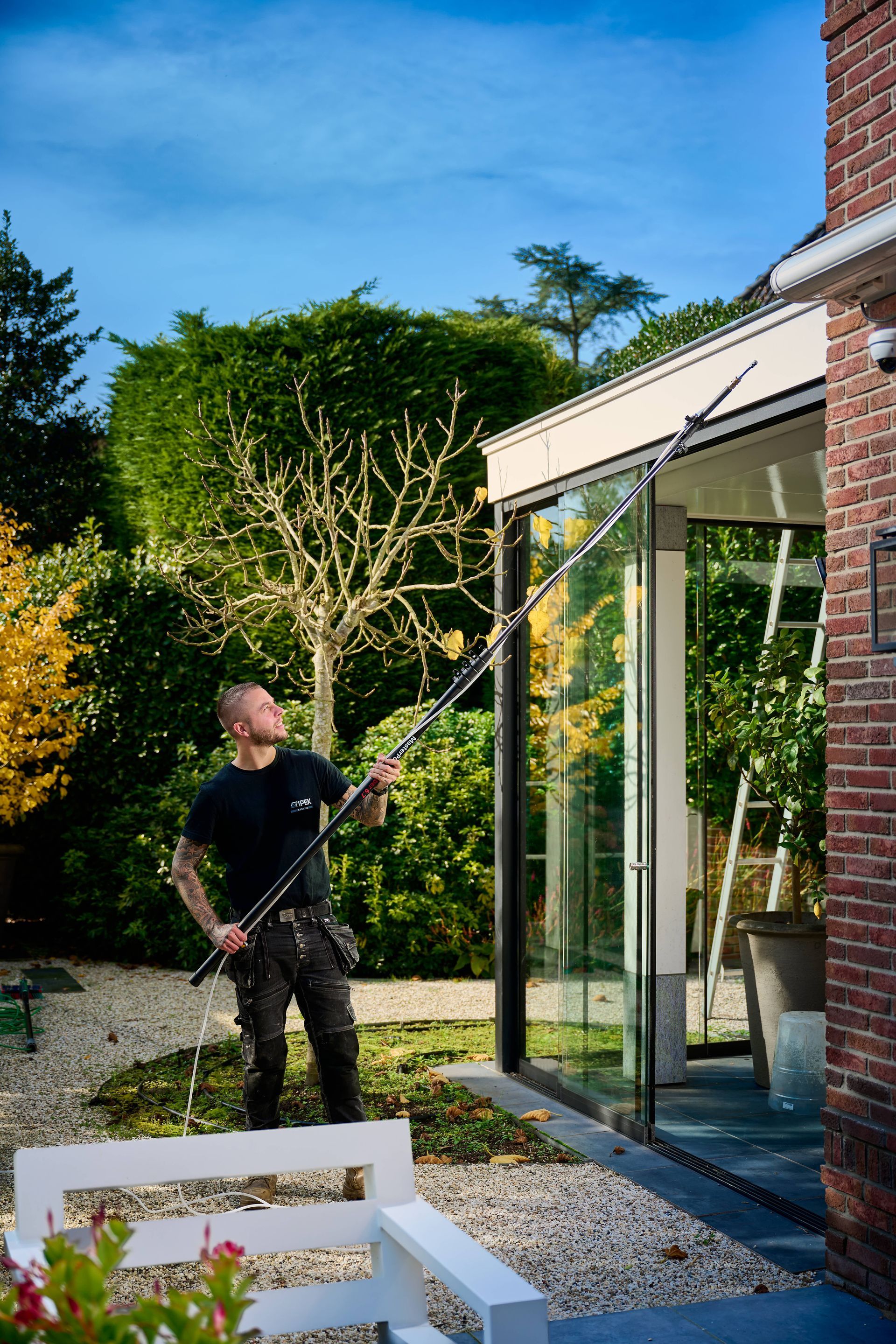 Een glazenwasser gebruikt een telescoopsteel om de glazen wanden van een gebouw te wassen, met op de achtergrond een weelderige groene tuin.