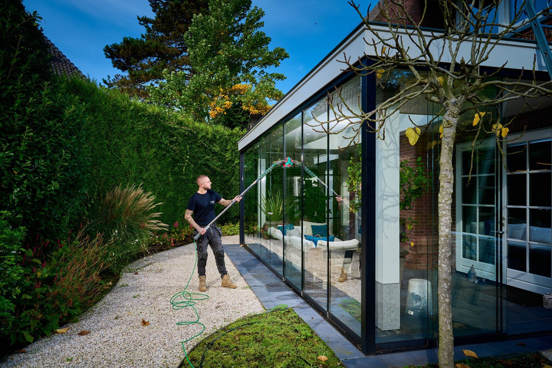 Een man maakt met een gereedschap met lange steel glazen ramen schoon in een tuin.