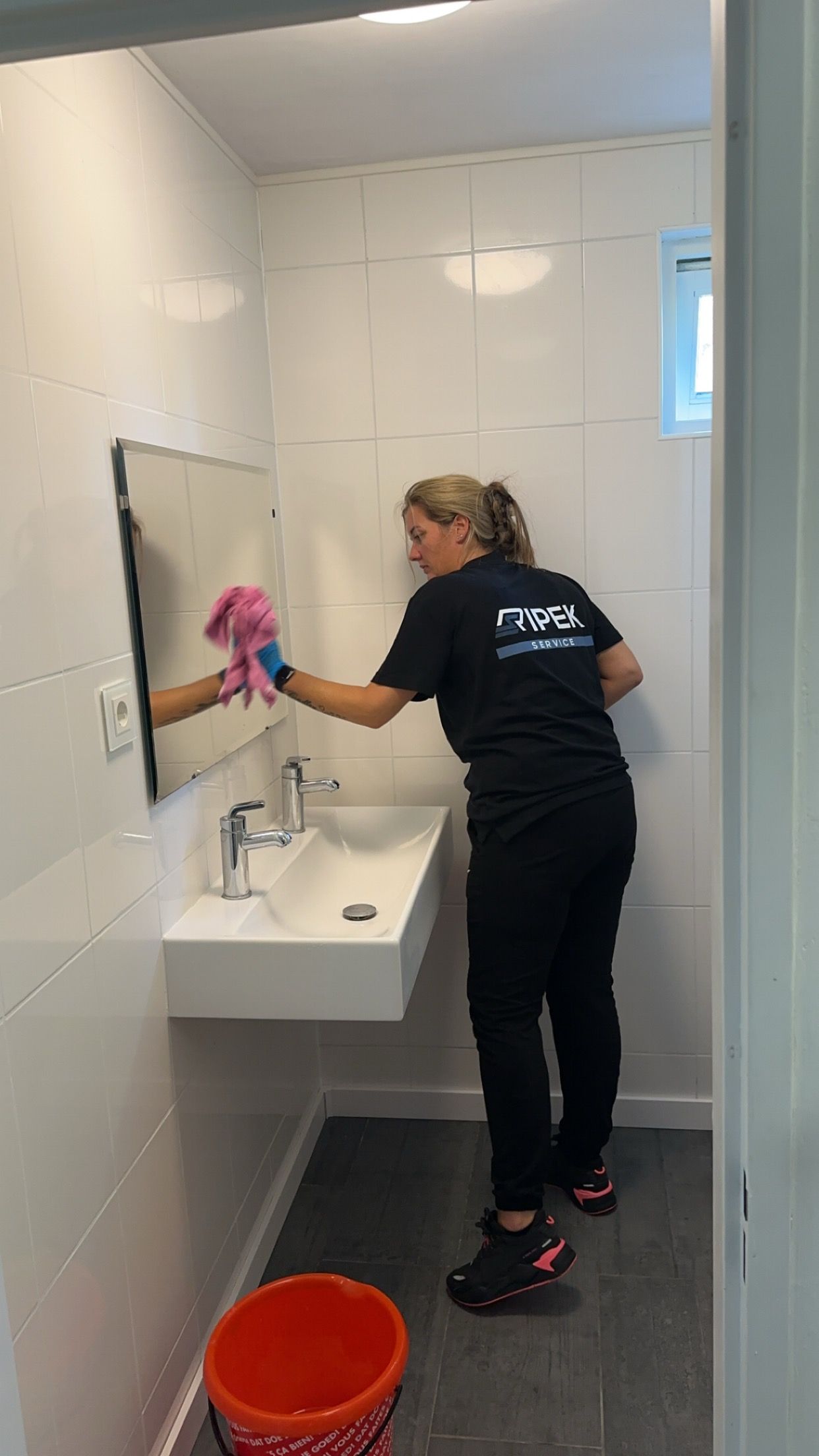 Iemand maakt een badkamerspiegel schoon met een roze doek, terwijl er een emmer met schoonmaakmiddel in de buurt staat.