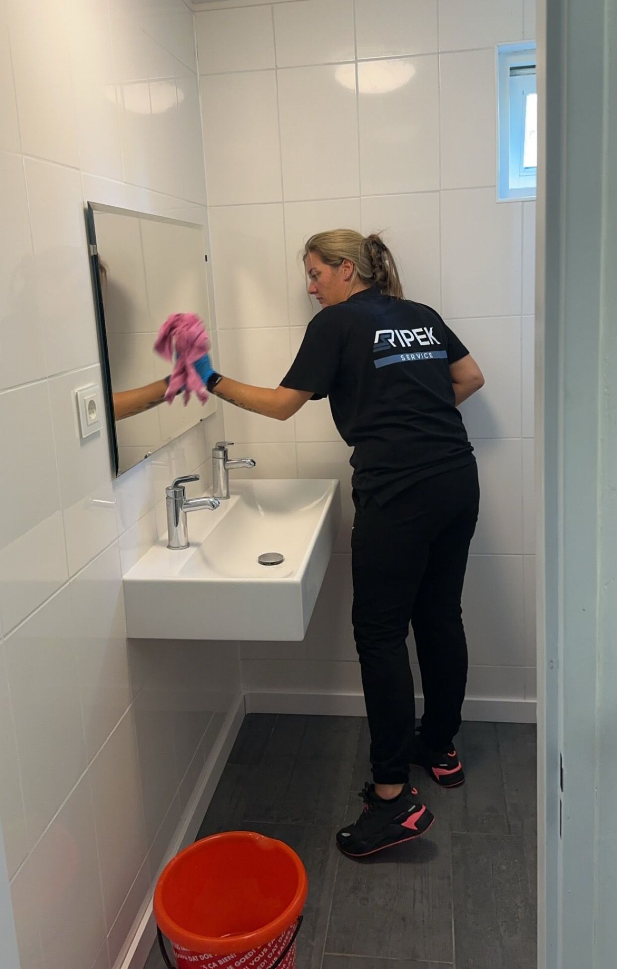 Persoon in zwart maakt een badkamerspiegel schoon met een roze doek; wastafel en emmer zijn zichtbaar.