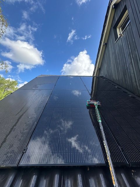 Zonnepanelen op een dak worden schoongemaakt met een borstel met lange steel, onder een blauwe hemel met wolken.