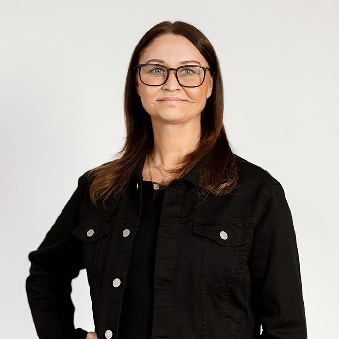 A woman wearing glasses and a black jacket is standing in front of a white background.