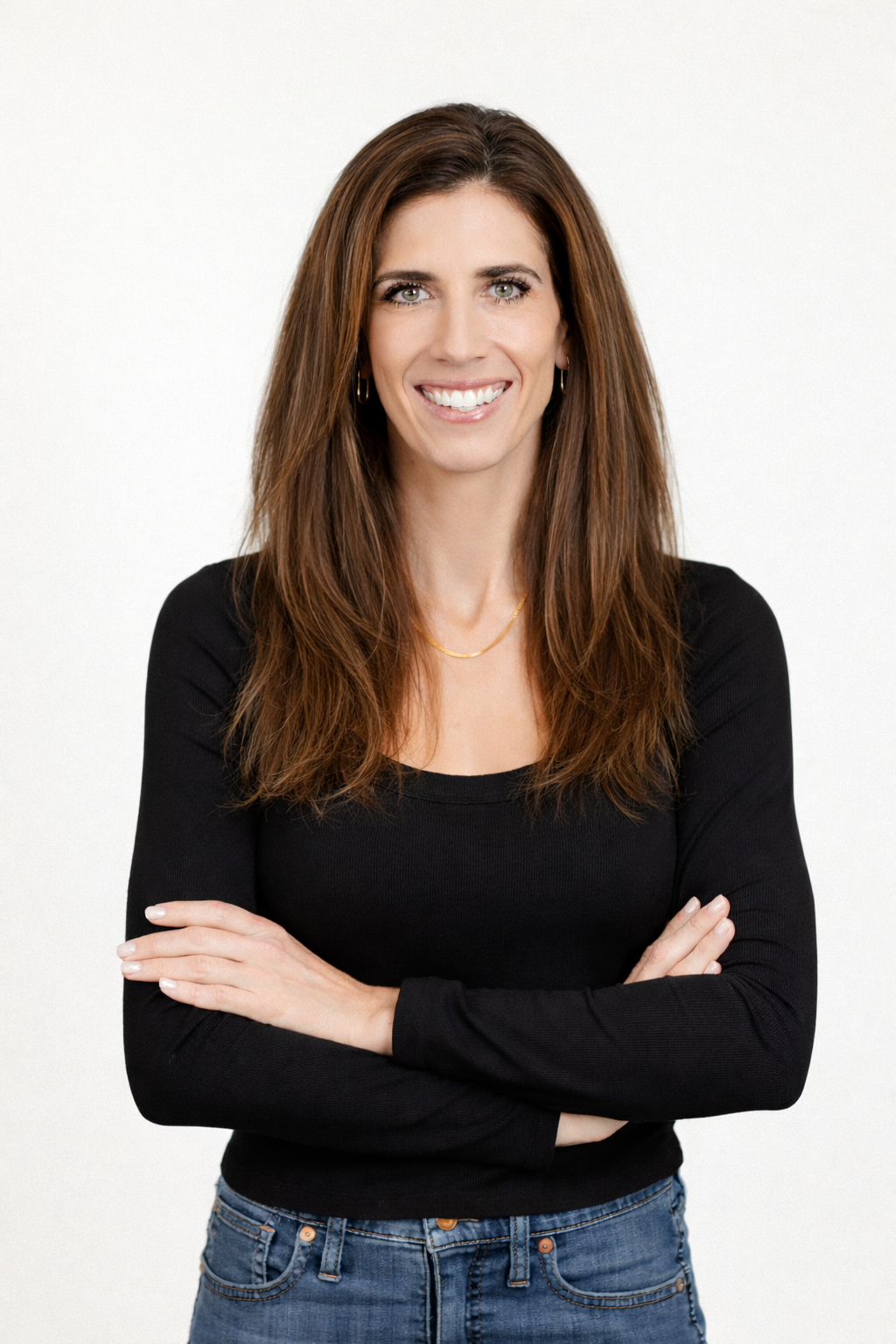 Woman standing with arms crossed, smiling, in a black top and jeans against a white background