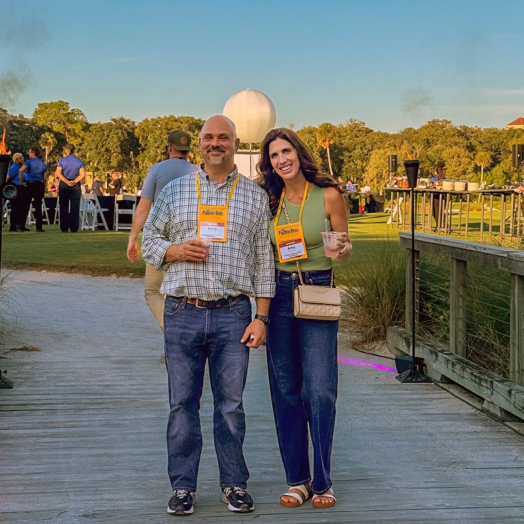 Two people stand on a wooden bridge at sunset, smiling and holding drinks, with water and trees behind them